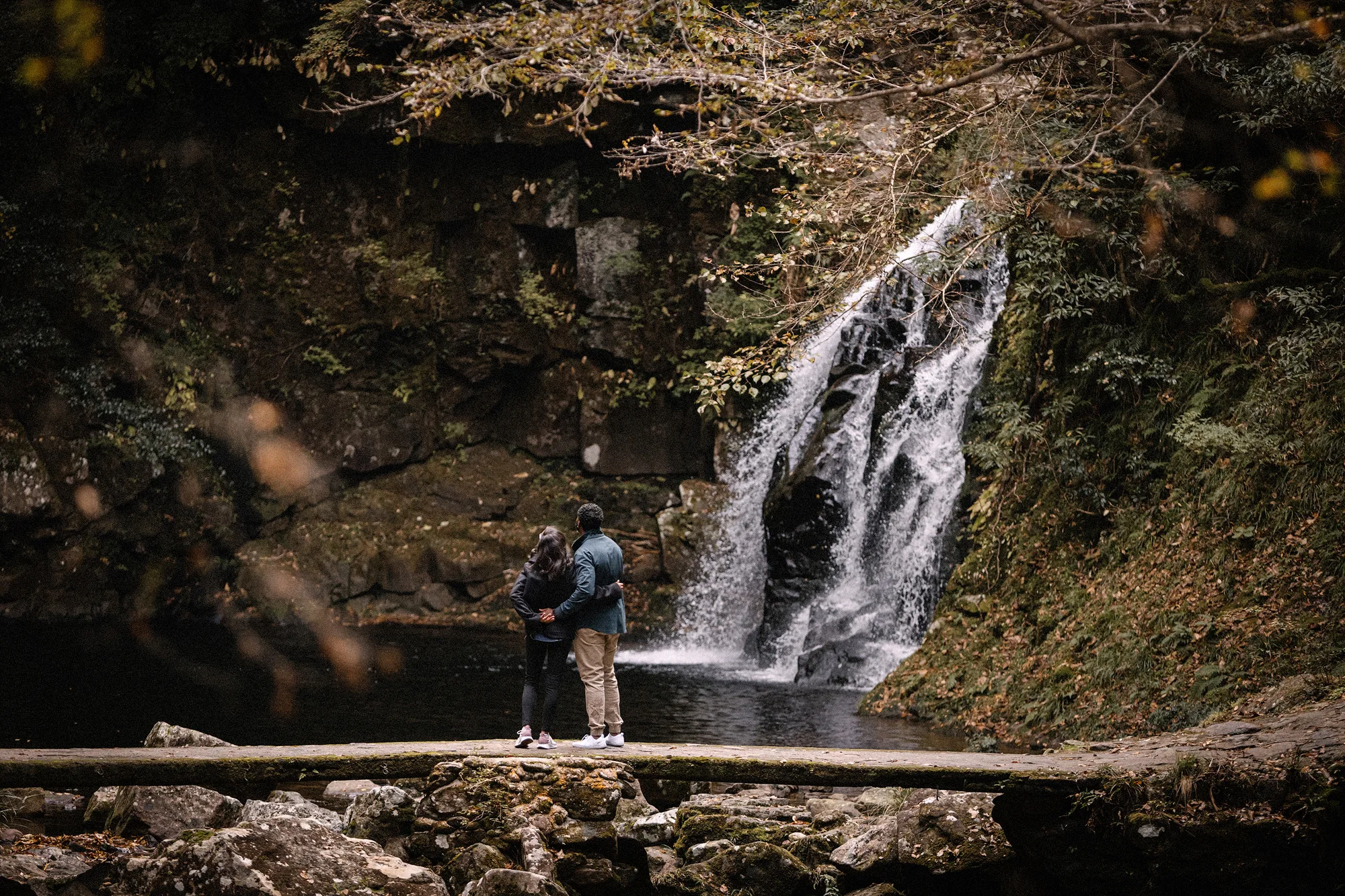 A couple stand below a waterfall in a mossy gorge at Akame 48 Falls.