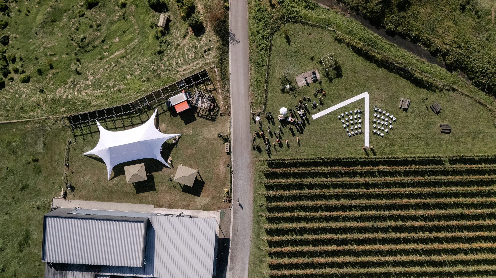 From above, a white aisle and rows of chairs set out on the lawn beside the vineyard at Akiu Winery, a tent and the winery roof nearby.