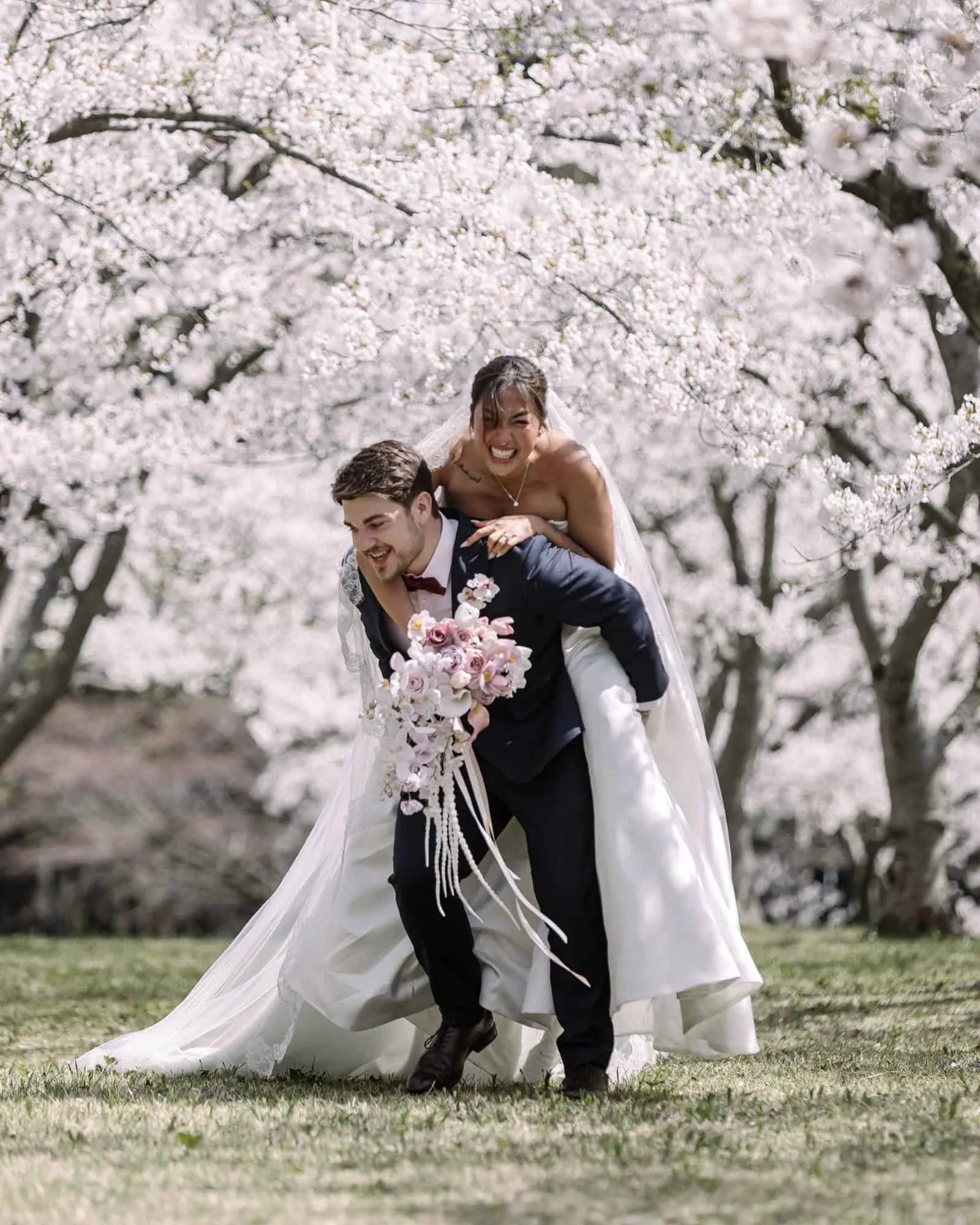 The groom gives the bride a piggyback under the cherry blossoms, her bouquet in one hand and a wide smile on her face.