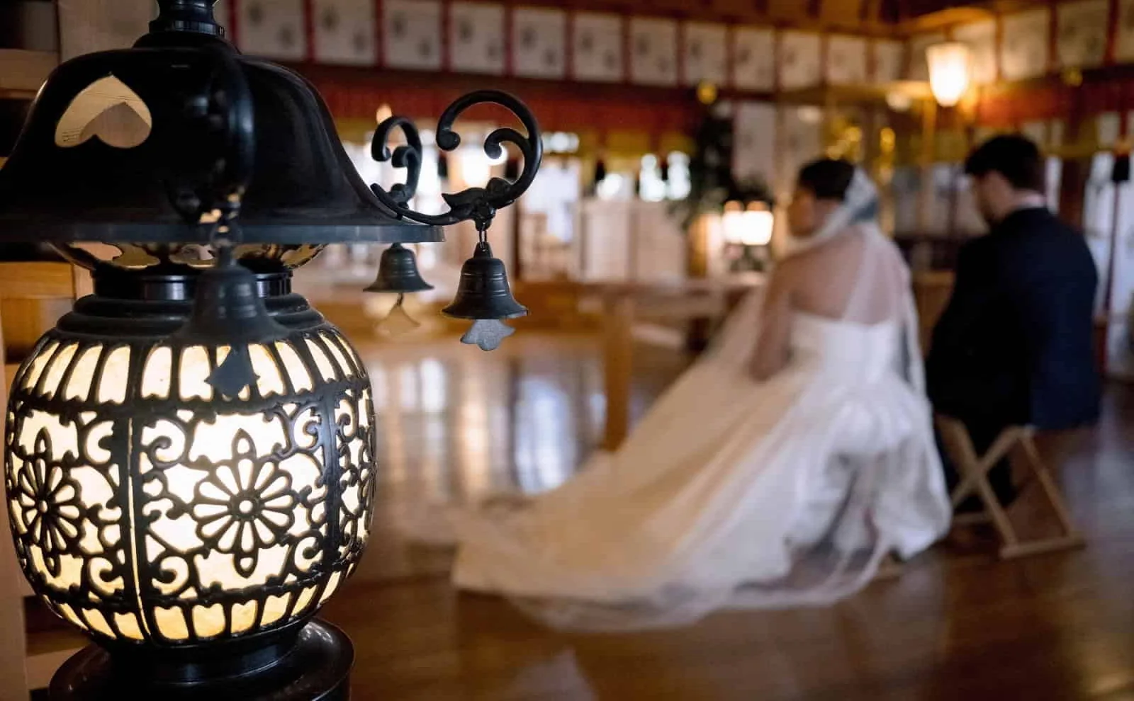 The bride in her gown and the groom sit in the hall at Takayama Inari Shrine during their blessing, a lantern glowing in the foreground.