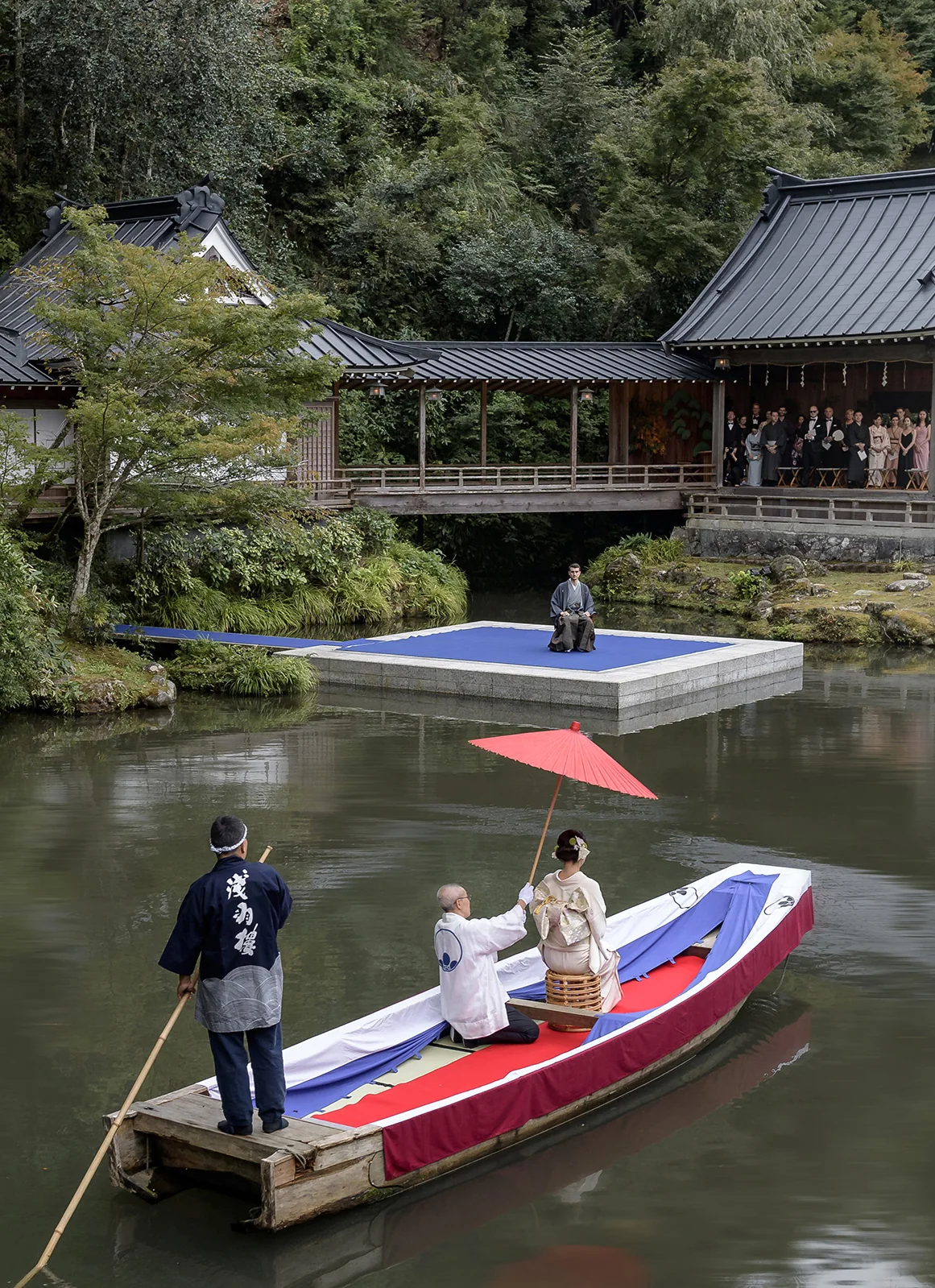 A bride in kimono crosses the pond by boat beneath a red parasol toward the floating Noh stage at Asaba Ryokan, guests watching from the veranda.