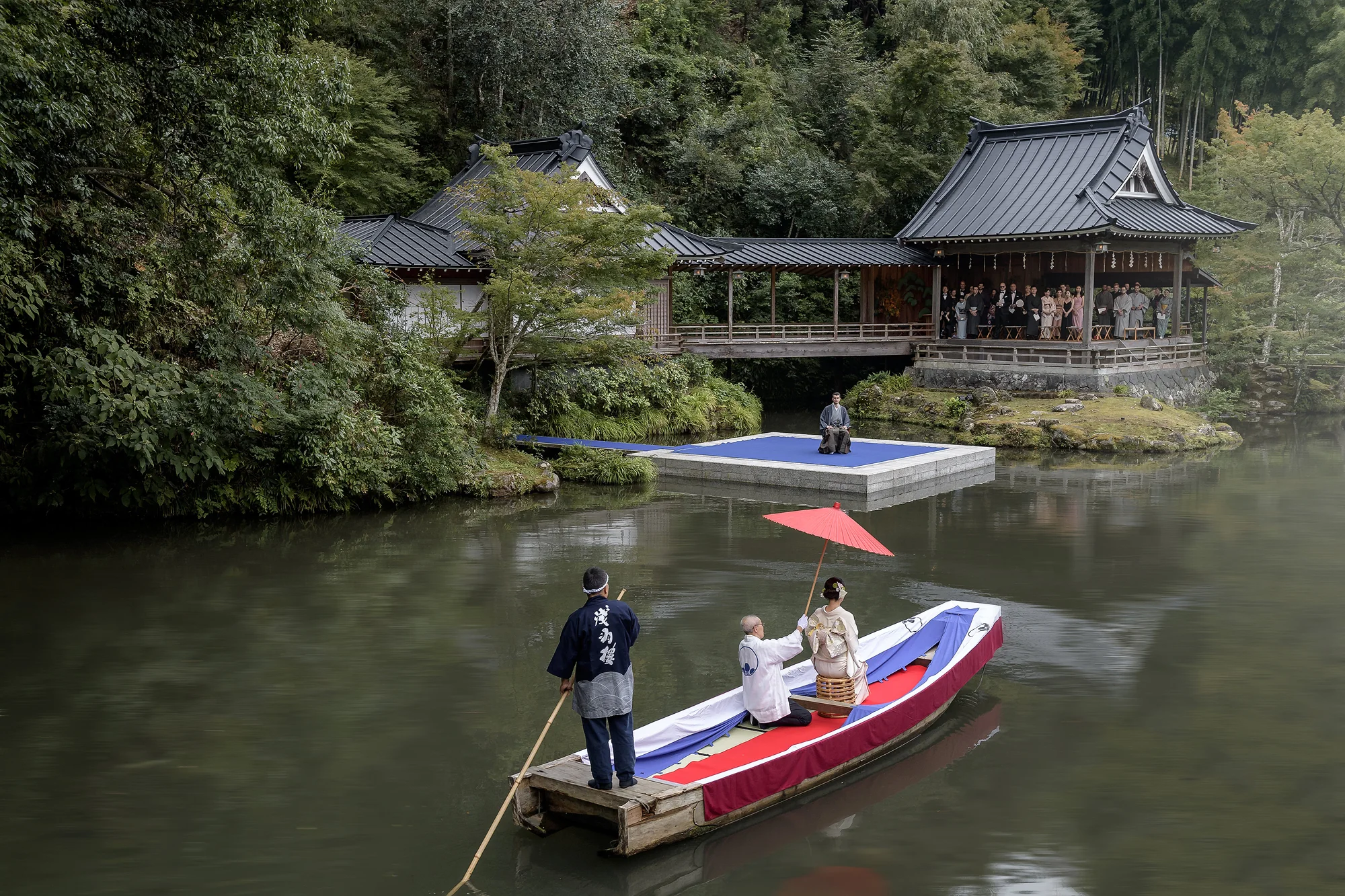 A boatman poles the couple across the pond toward the floating Noh stage at Asaba Ryokan, a red umbrella held over them, guests watching from the hall.