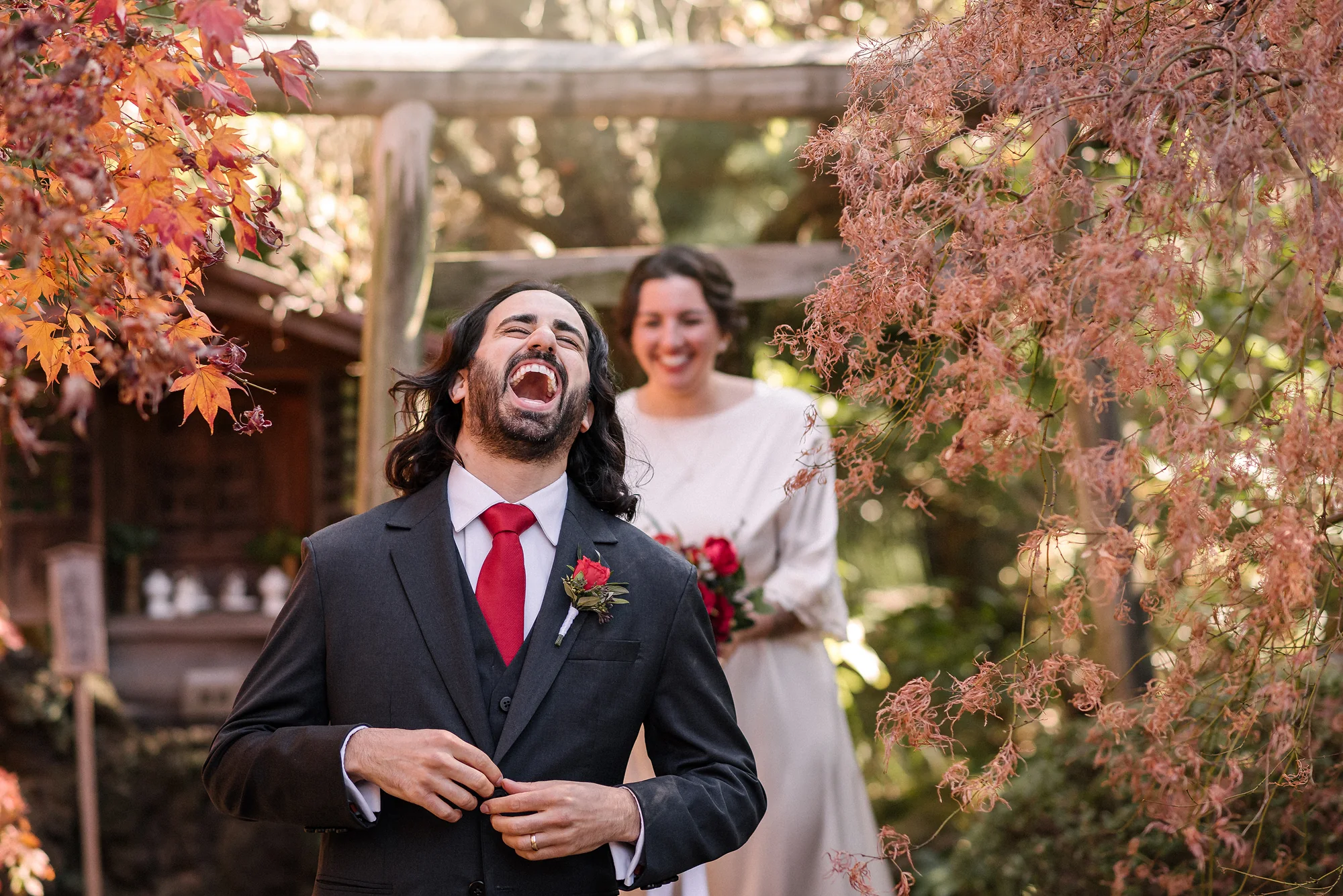 The groom throws his head back laughing as the bride smiles beside him among autumn leaves.