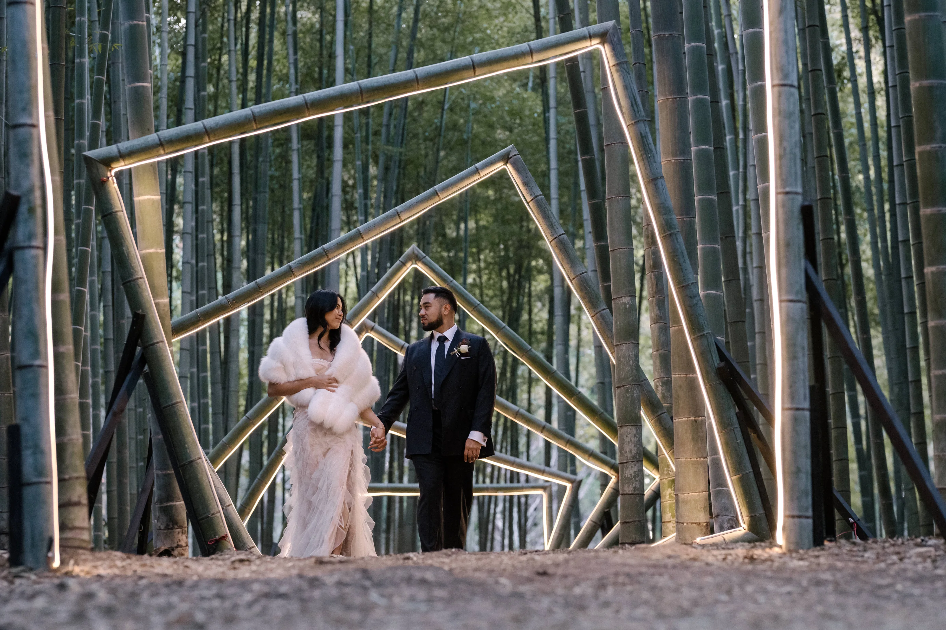The couple walk hand in hand through nested square frames of bamboo edged with light in a Tochigi bamboo forest, her white fur stole around her.