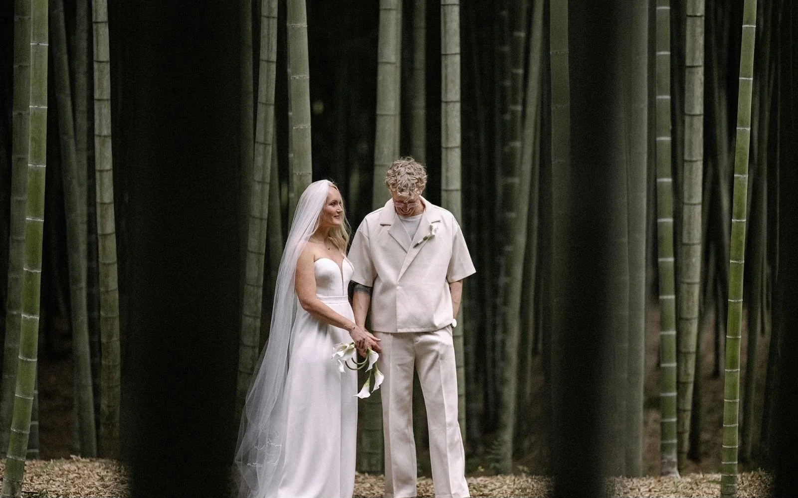 The bride in a long veil and the groom stand hand in hand among tall bamboo.