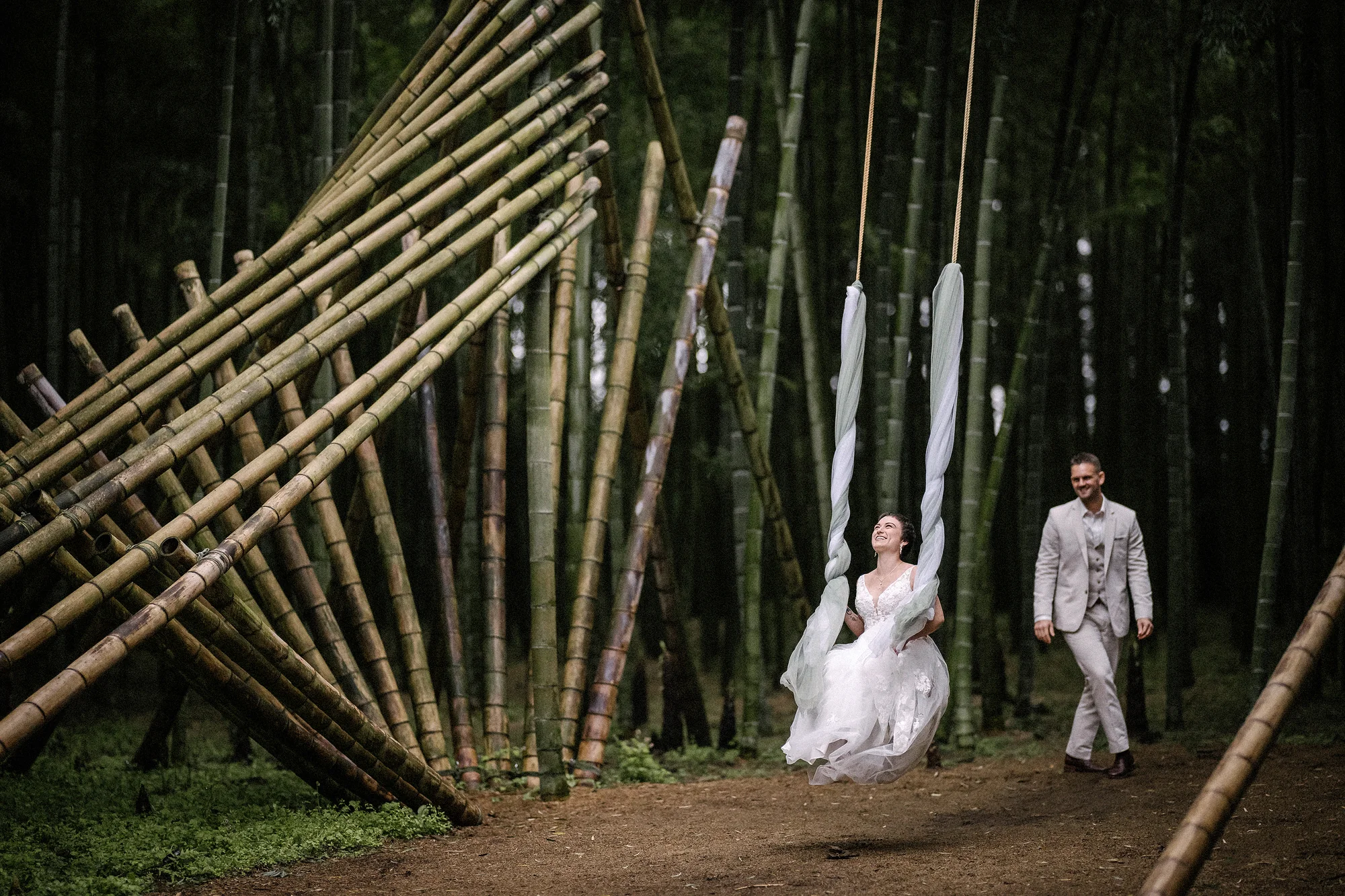 The bride swings on a rope swing among leaning bamboo poles as the groom looks on.