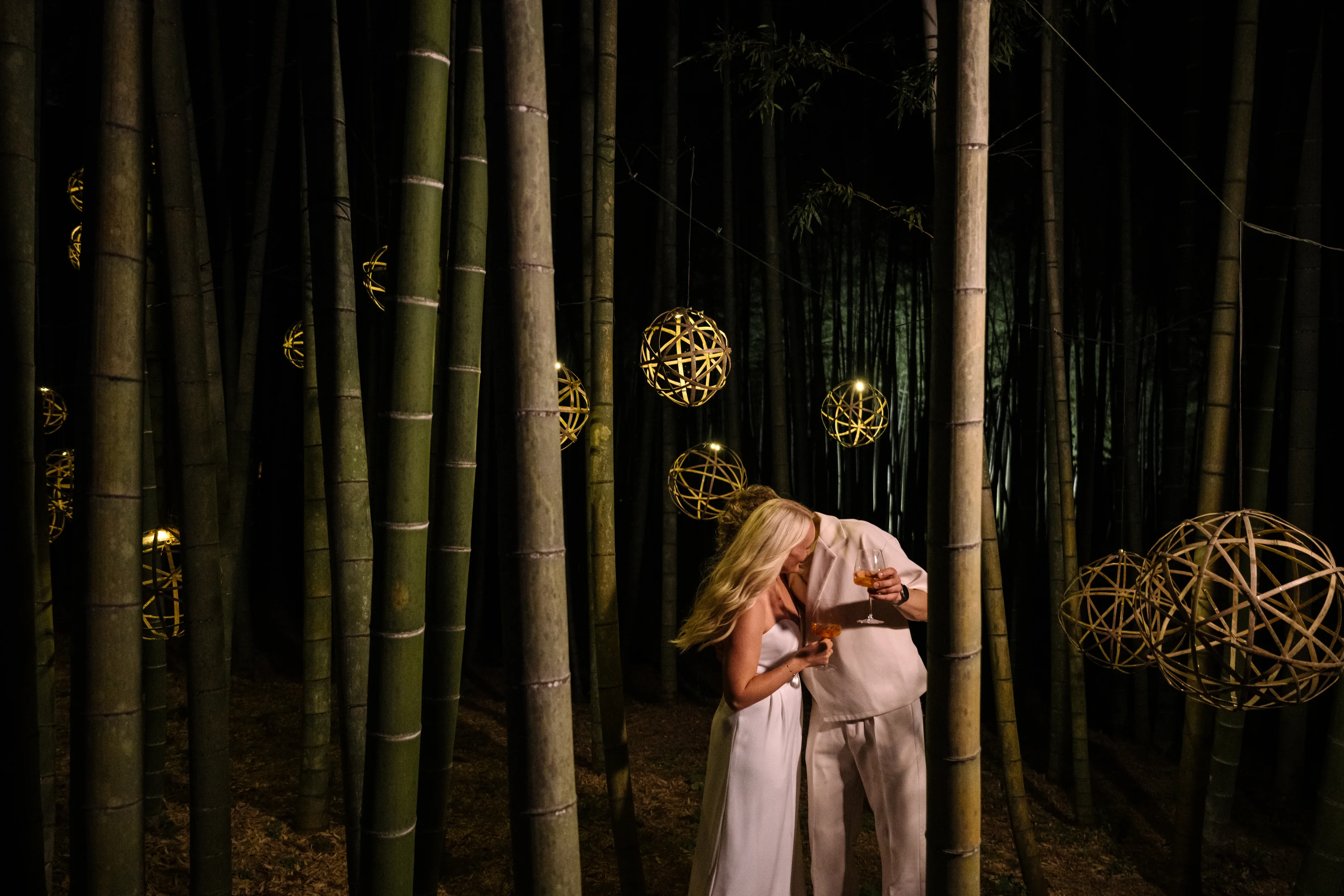 A couple share drinks beneath lanterns in a bamboo forest.