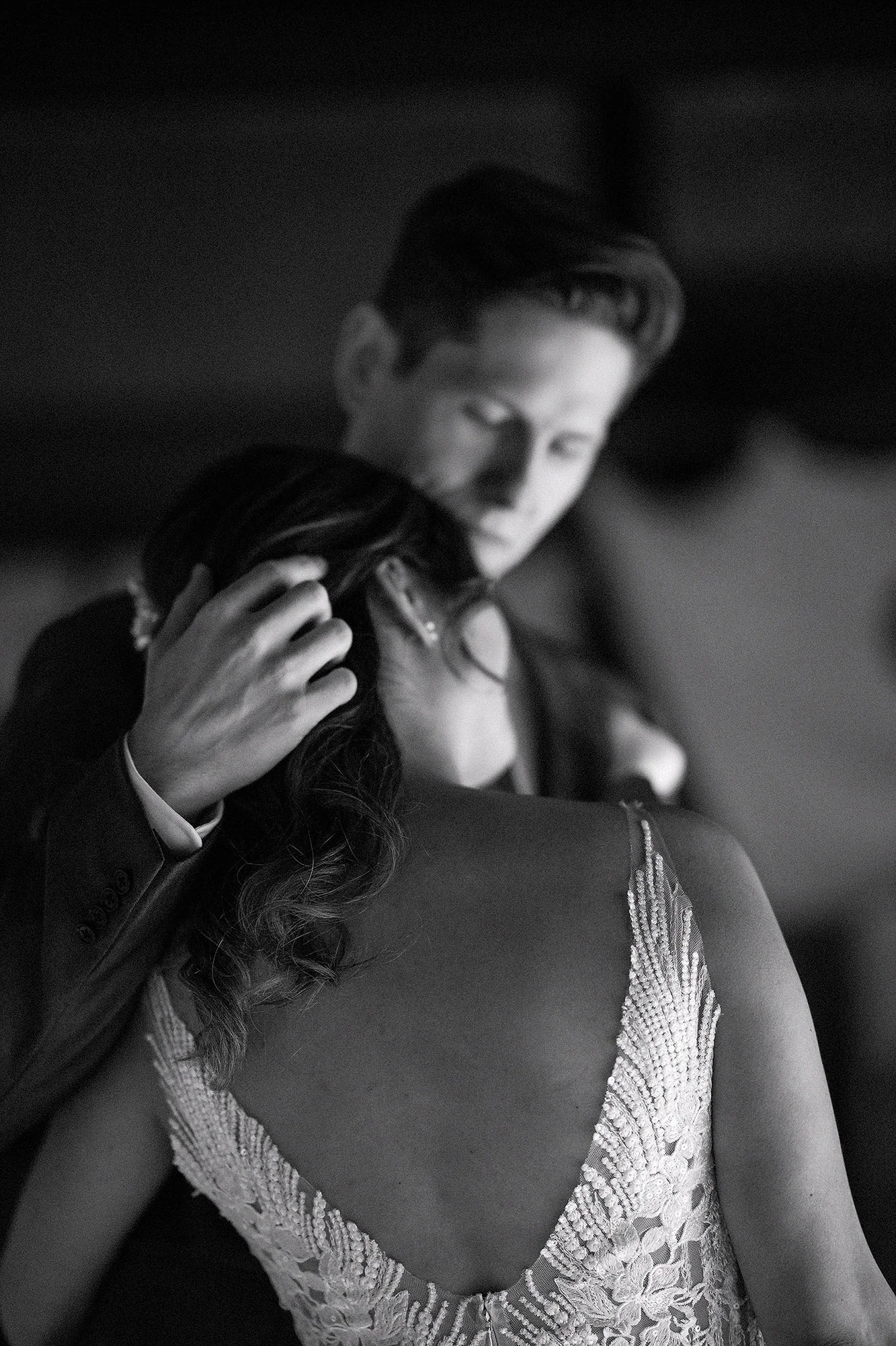 In black and white, the groom holds the bride close during a slow dance, his hand in her hair, the lace back of her gown in the foreground.
