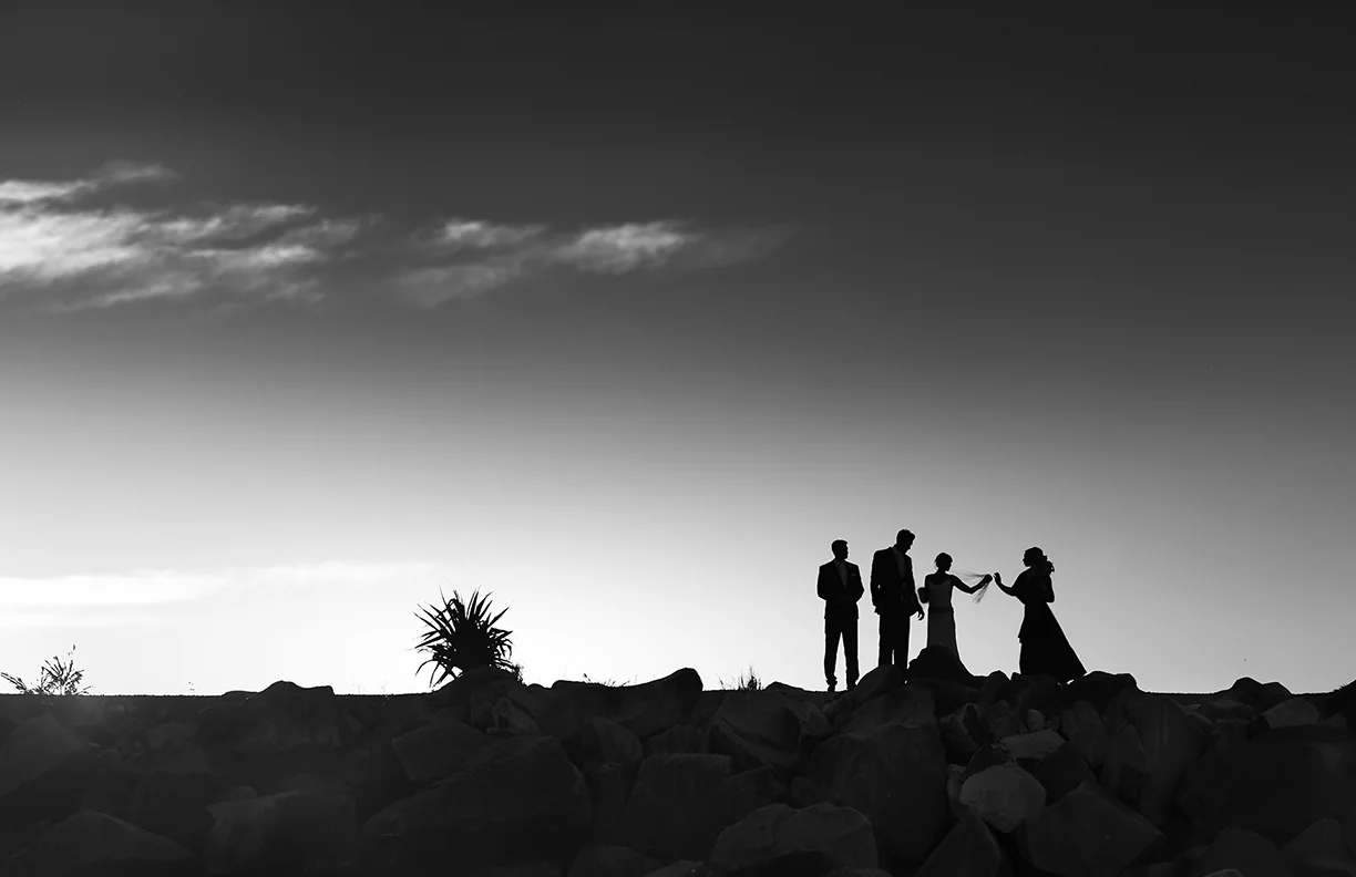In black and white, the bridal party stand in silhouette on a rocky headland under a wide evening sky in Noosa.