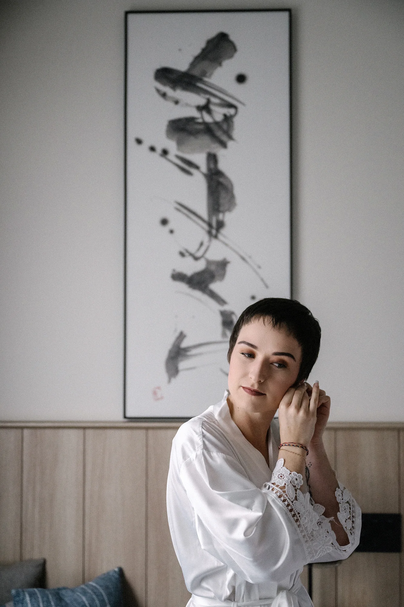 The bride fastens an earring in a white robe in Utsunomiya, a hanging scroll of calligraphy on the wall behind her.