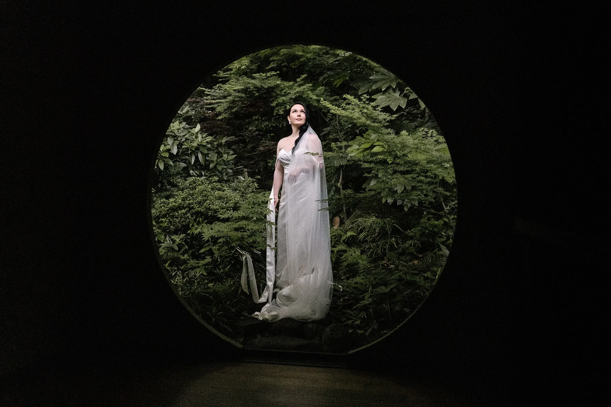 The bride stands framed by a round window onto a green garden.