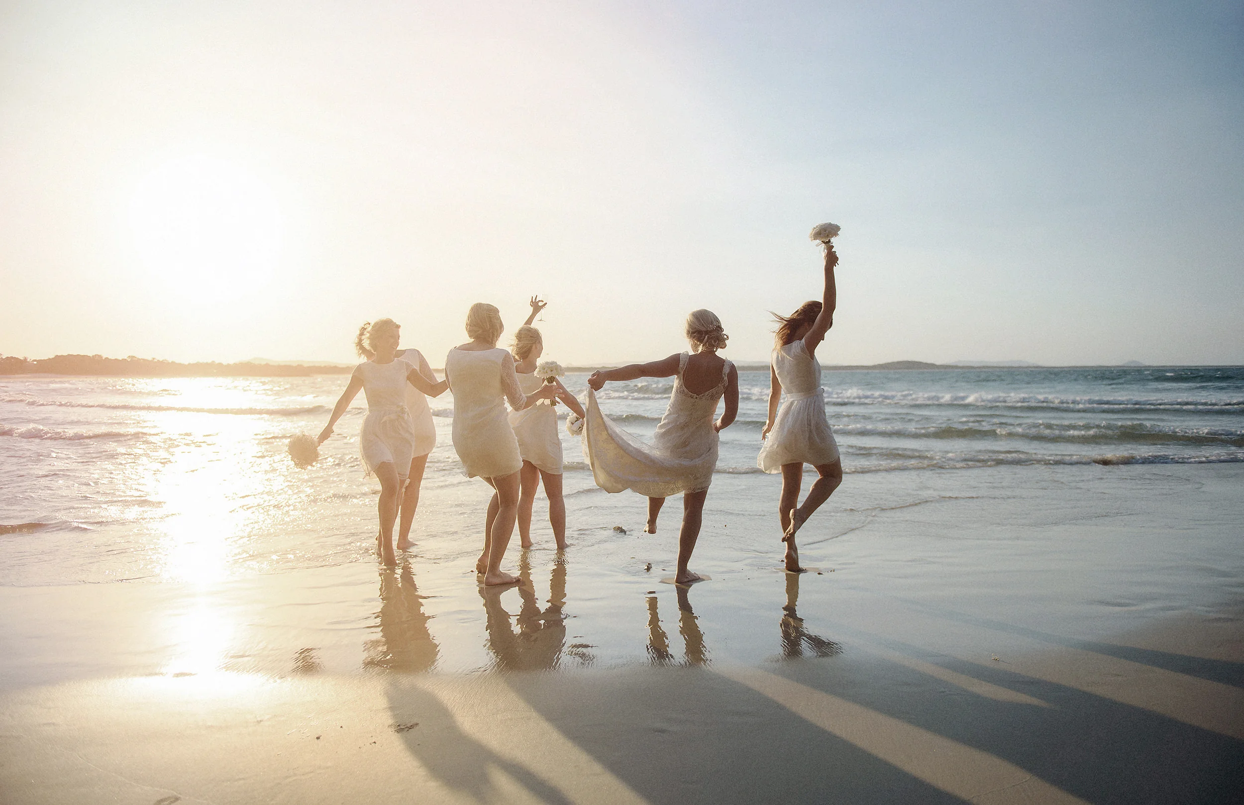 The bride and her bridesmaids run laughing along the water's edge into a low golden sun, bouquets held high.