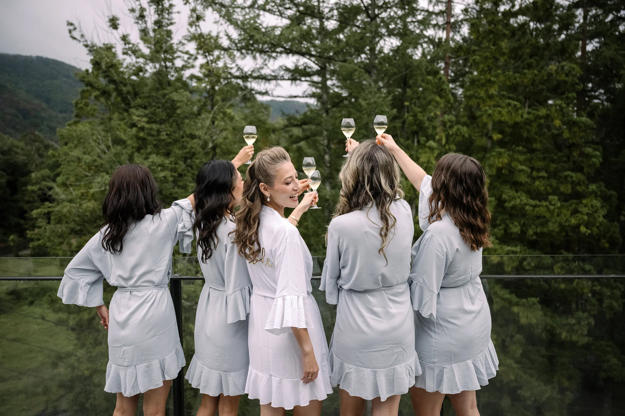 Bridesmaids in matching blue robes raise their glasses toward the mountains.