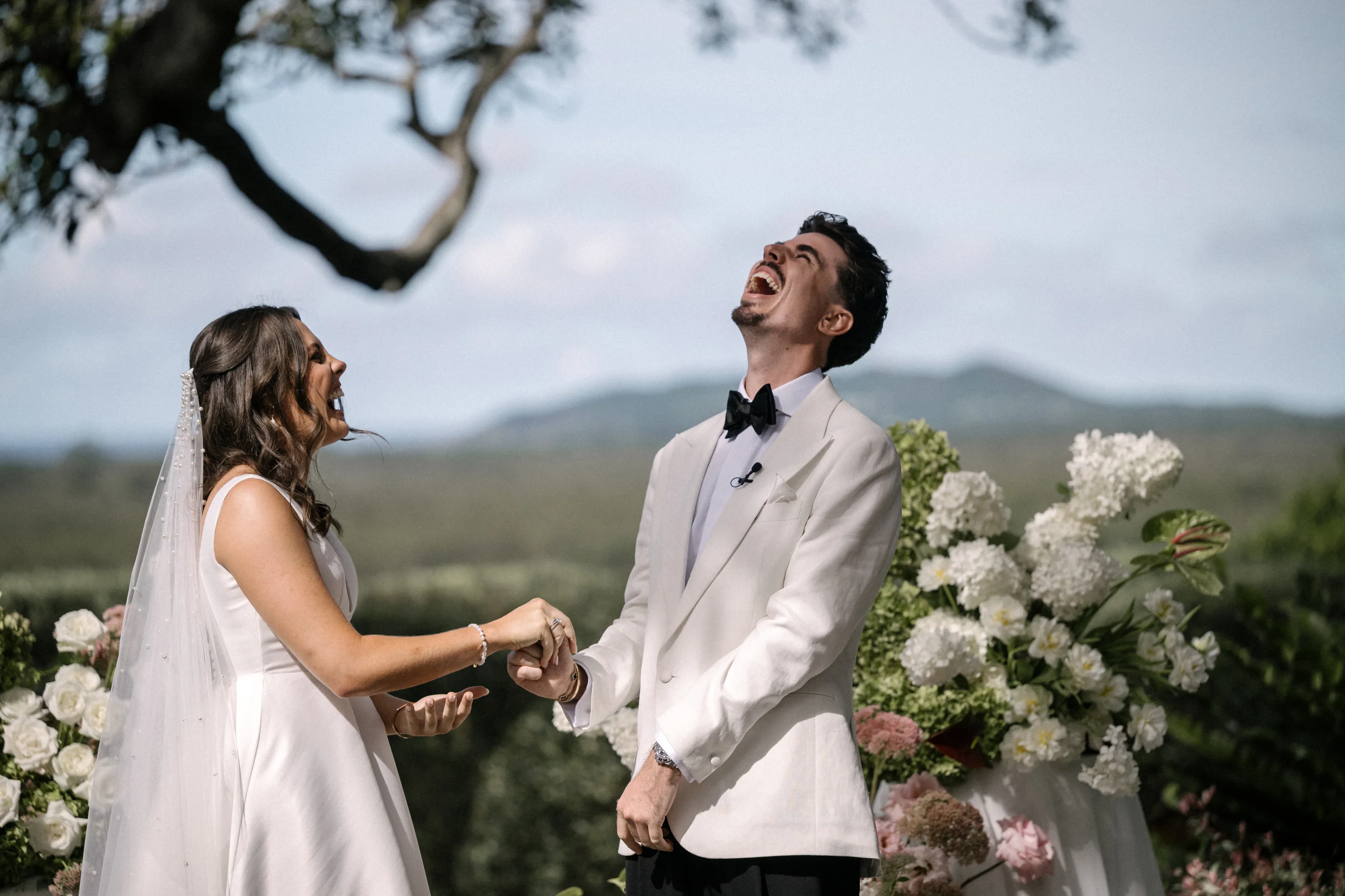 A groom throws his head back laughing as he and his bride hold hands at the ceremony.