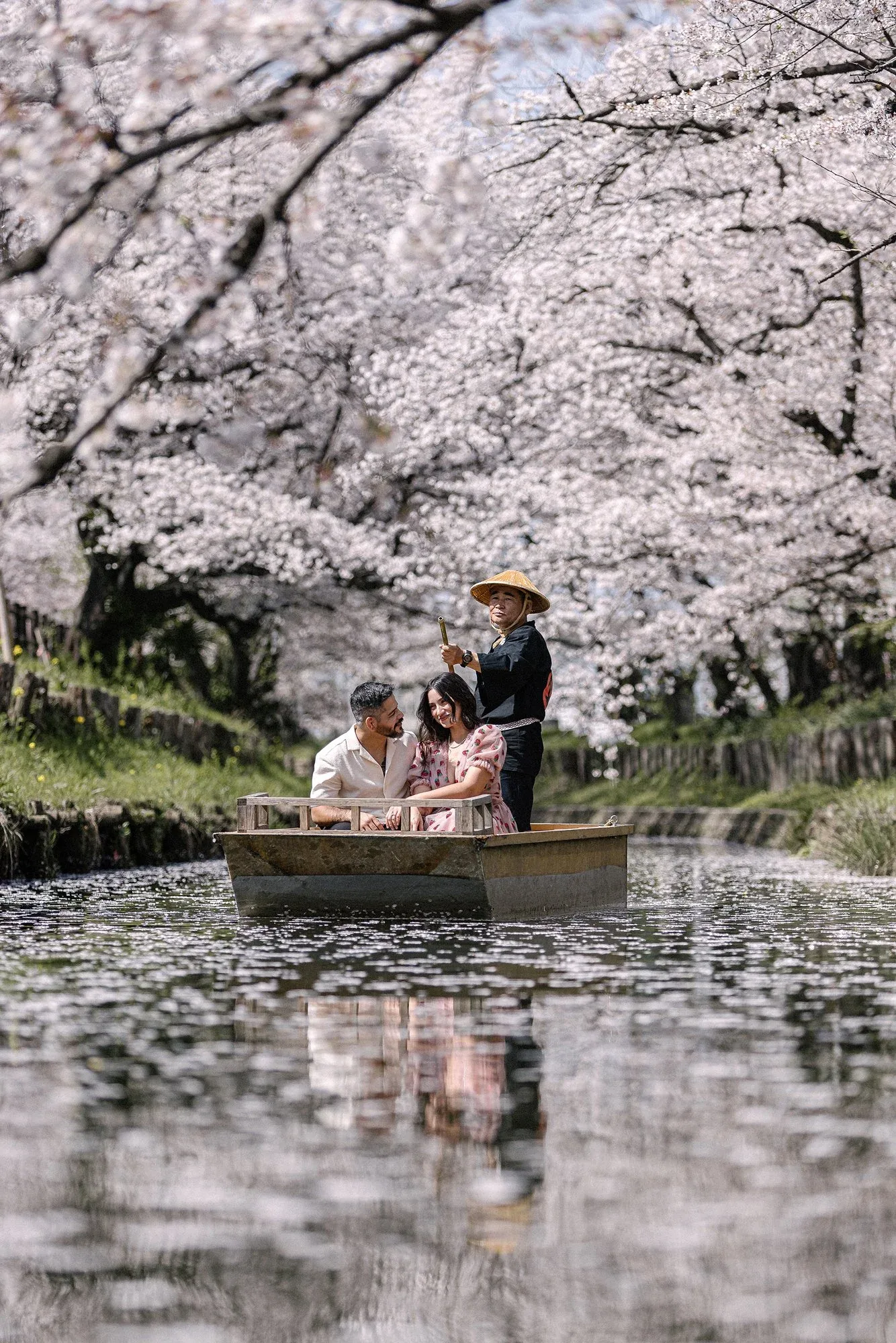 A boatman guides a couple along a canal in Kawagoe beneath cherry trees in full bloom, petals on the water.