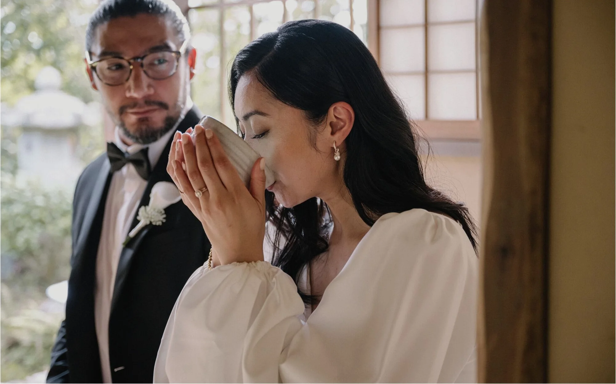 The bride and groom sip matcha together from tea bowls by a shoji window.