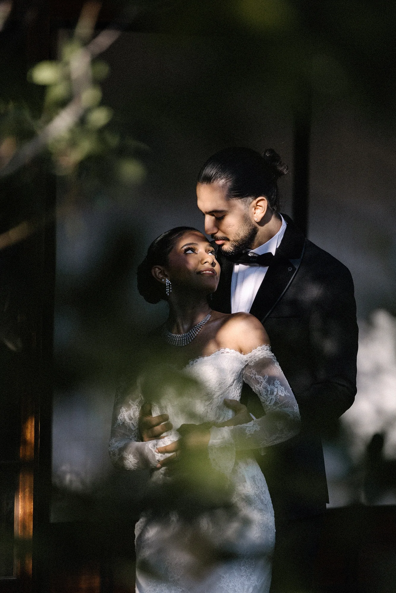 Seen through dark leaves, the groom holds the bride close outside a traditional Japanese house.