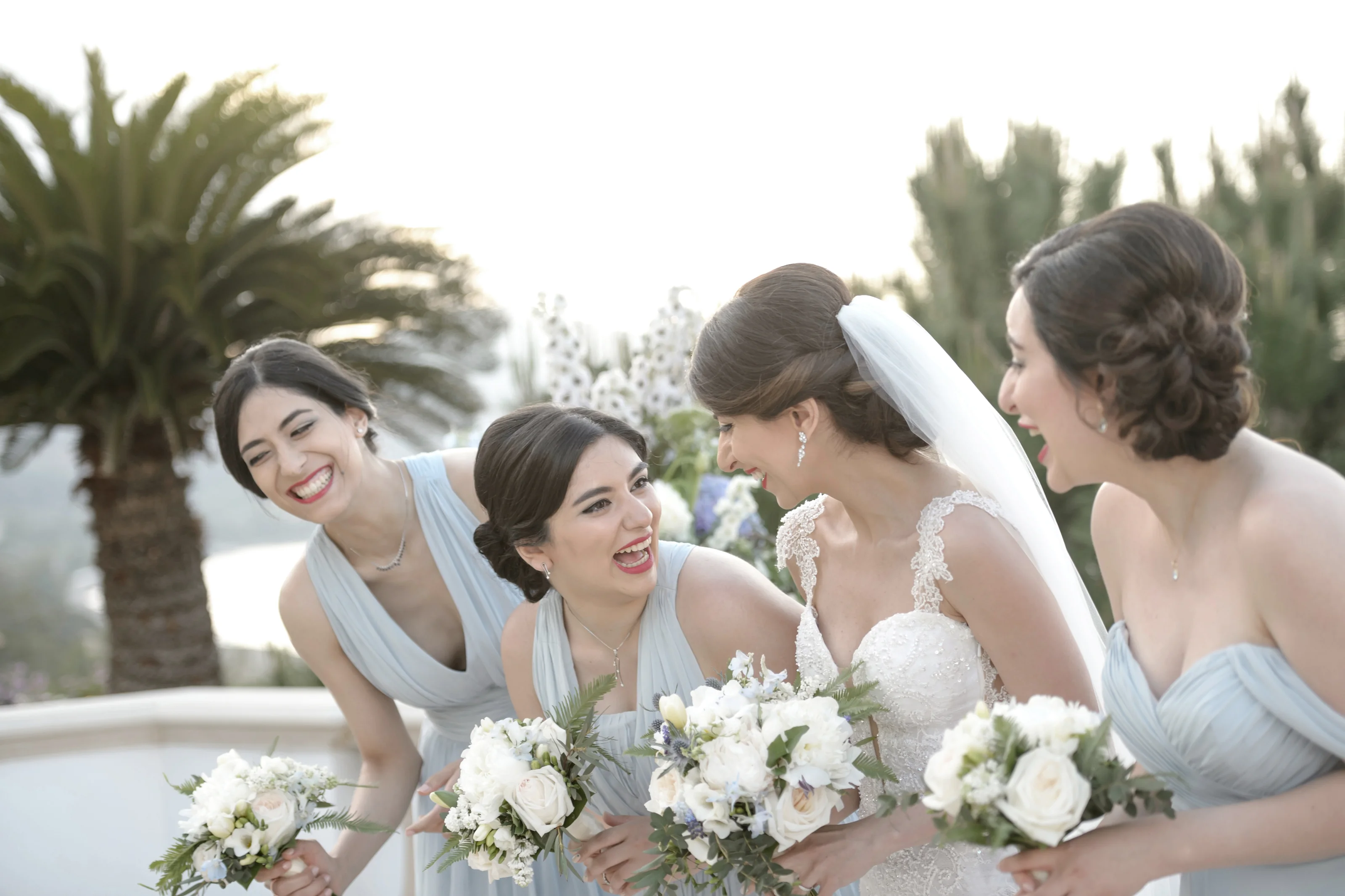 The bride and her bridesmaids lean in together, laughing, bouquets in hand, under palm trees in Cyprus.
