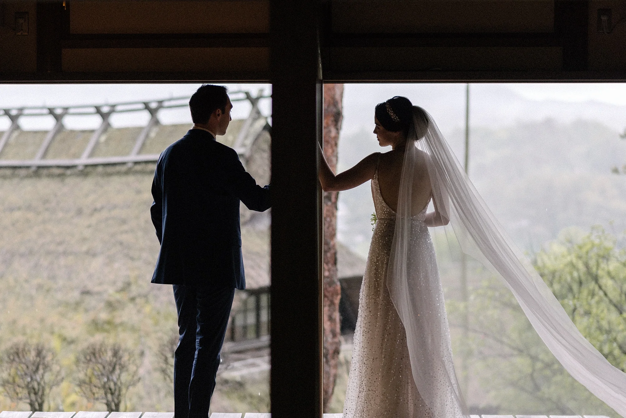 The groom waits at an open doorway as the bride looks out over thatched roofs and hills.