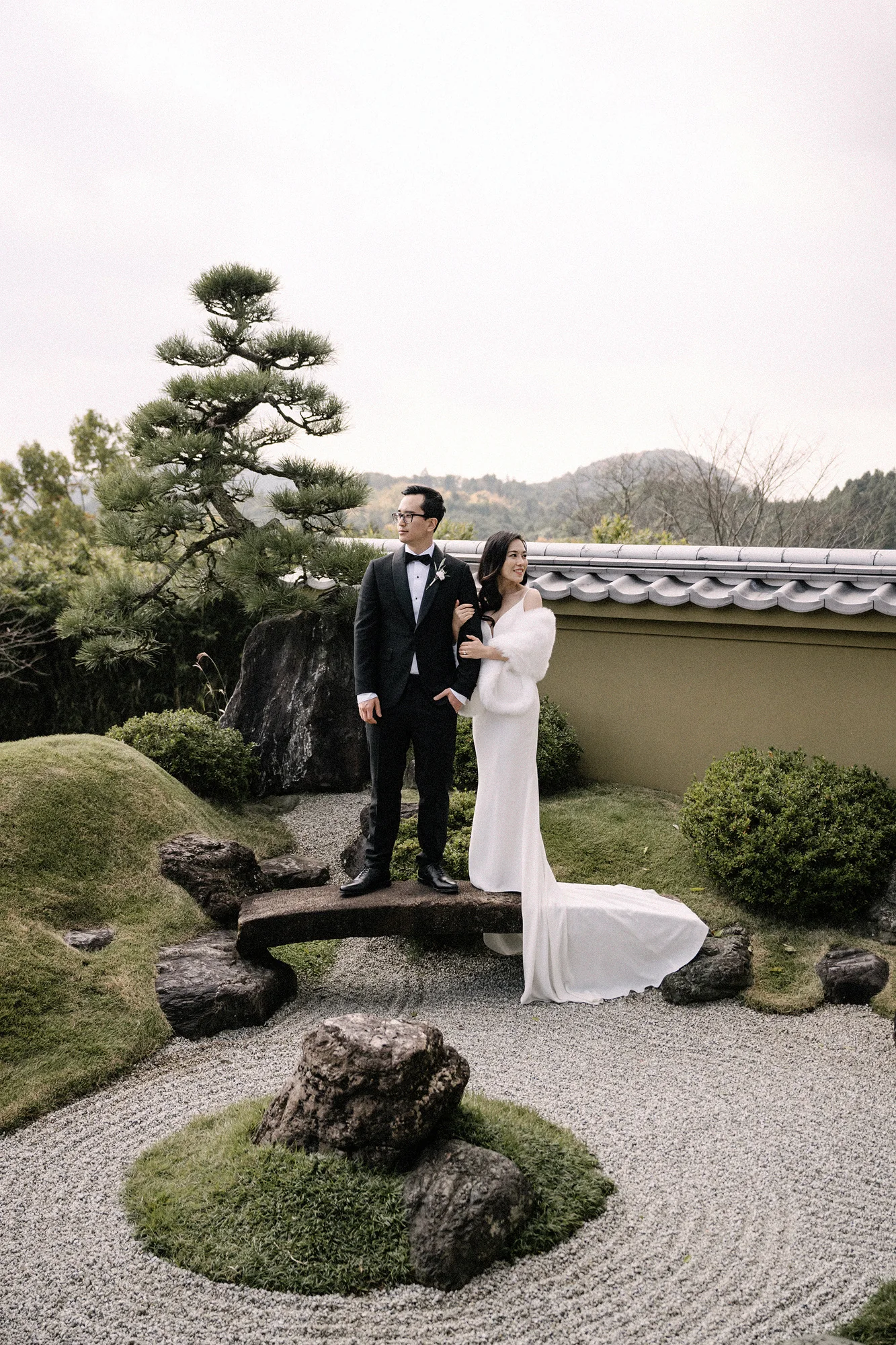 The couple stand on a stone bridge in a raked dry garden, pines and a white wall behind them.