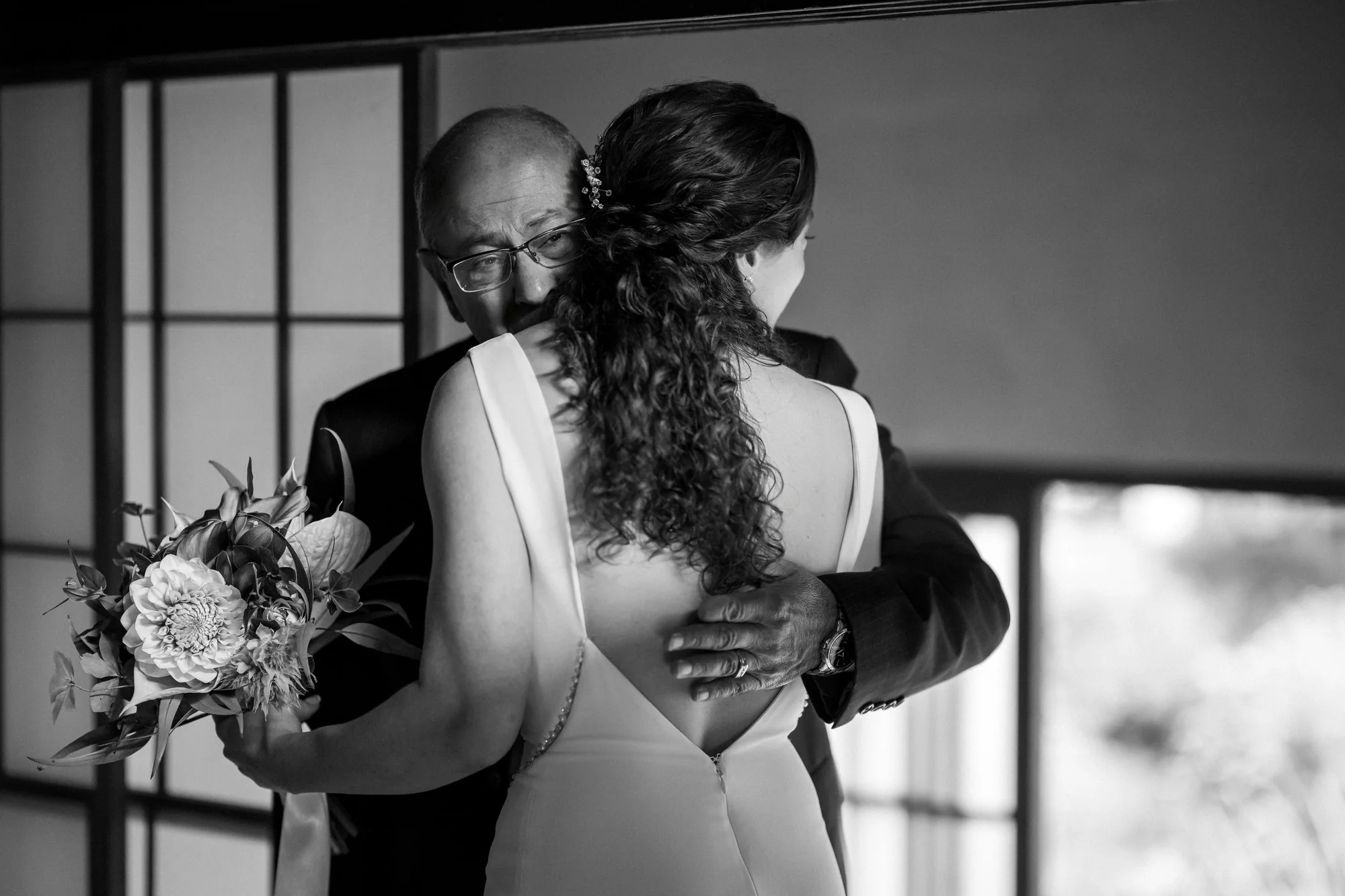 In black and white, a father holds his daughter close as he sees her in her wedding dress for the first time.