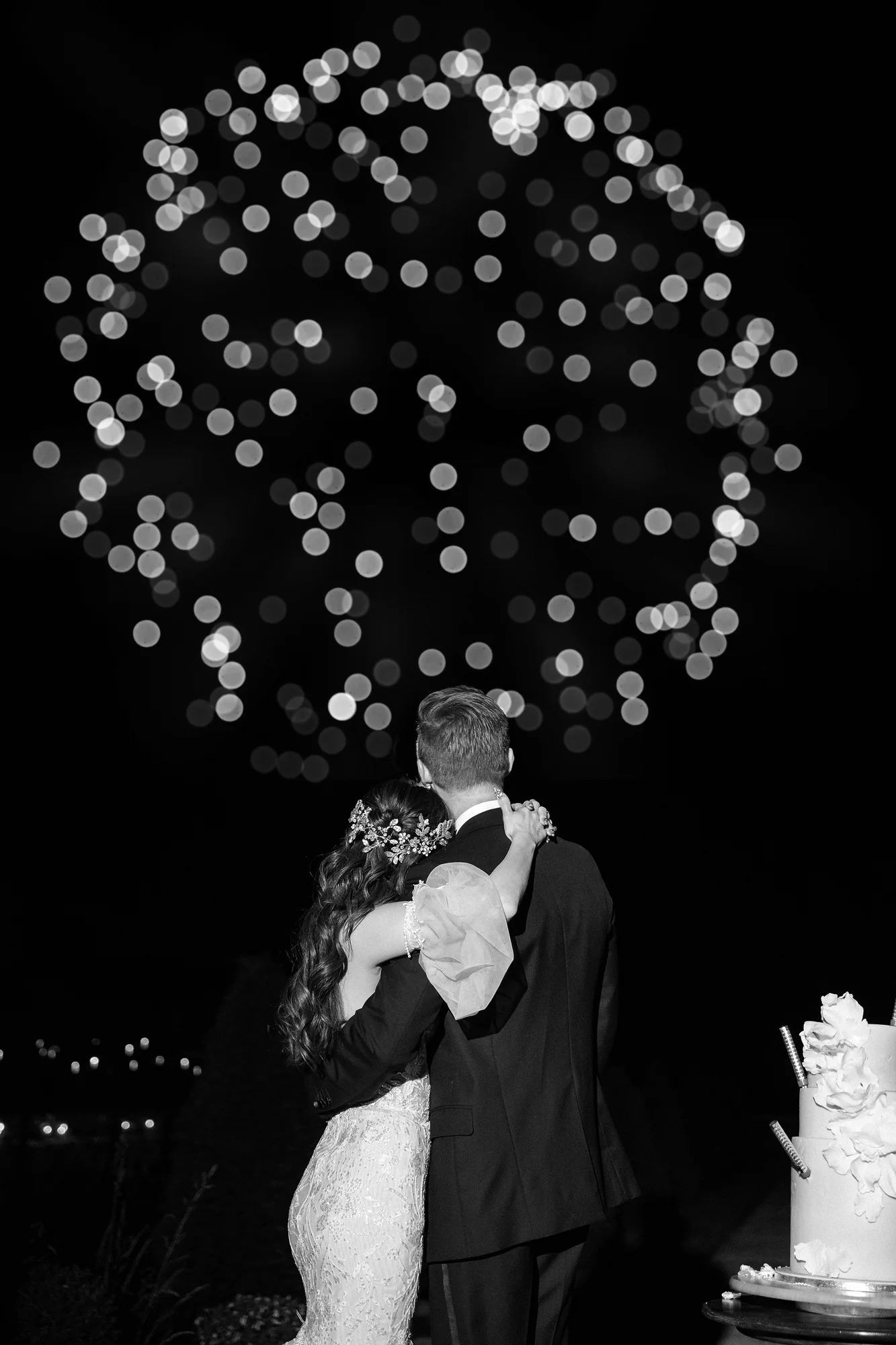 In black and white, the couple hold each other and watch fireworks burst above them at night, the wedding cake beside them.