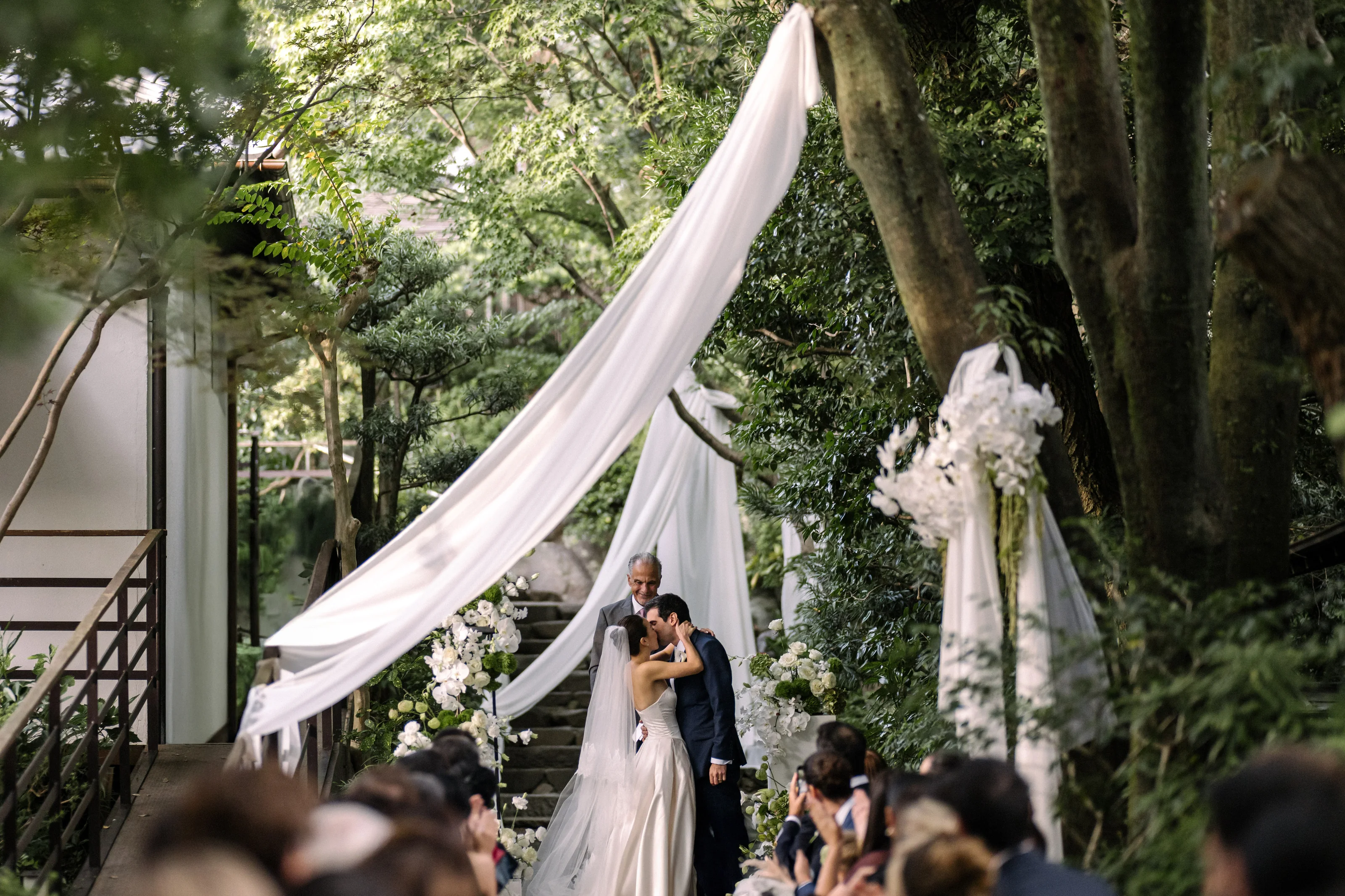 Bride and groom kissing beneath draped fabric and flowers on a tree-lined ceremony path in Kanagawa.