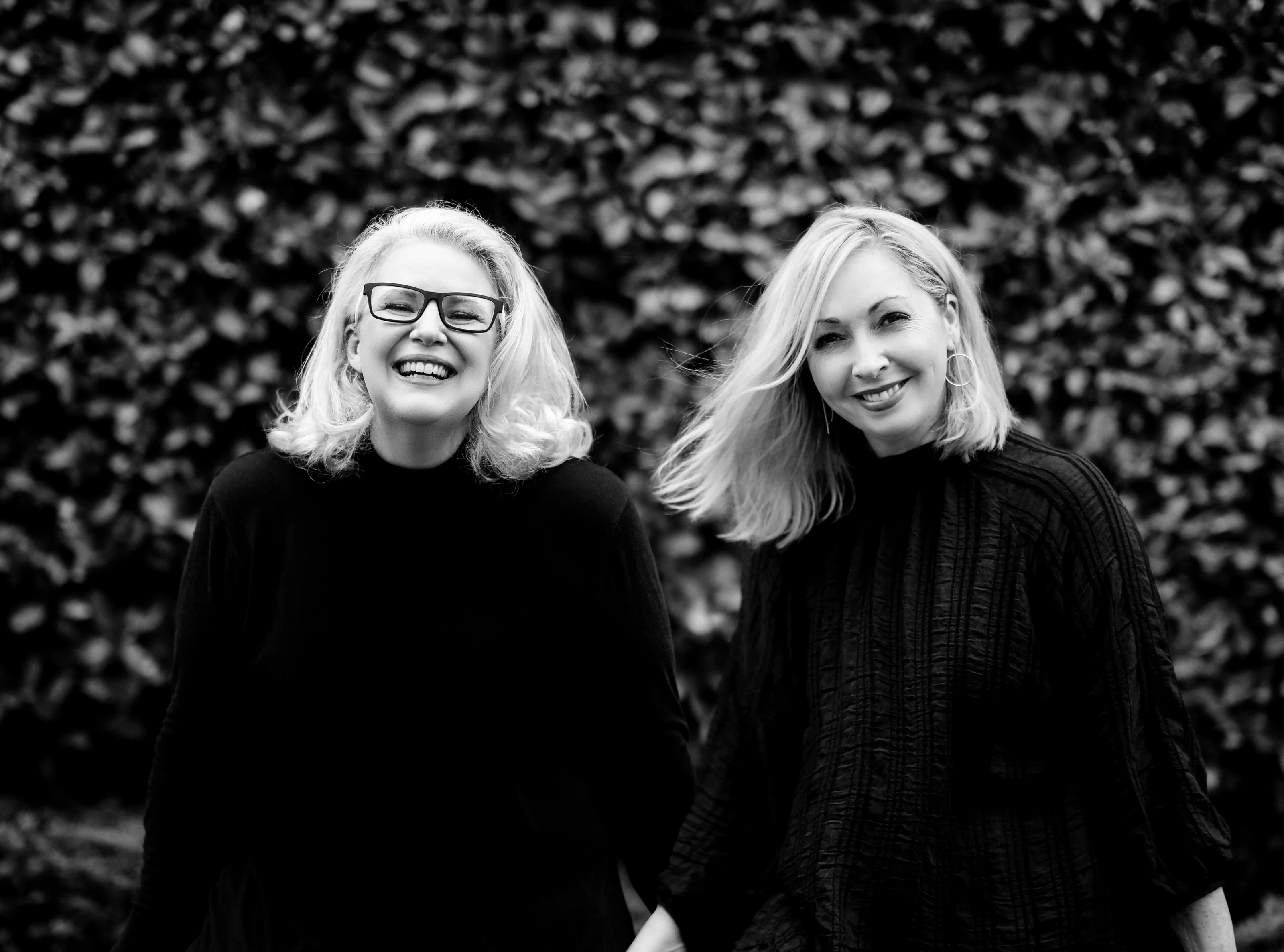In black and white, Tracey and Dee, the founders of 37 Frames, smile in front of a dark hedge.