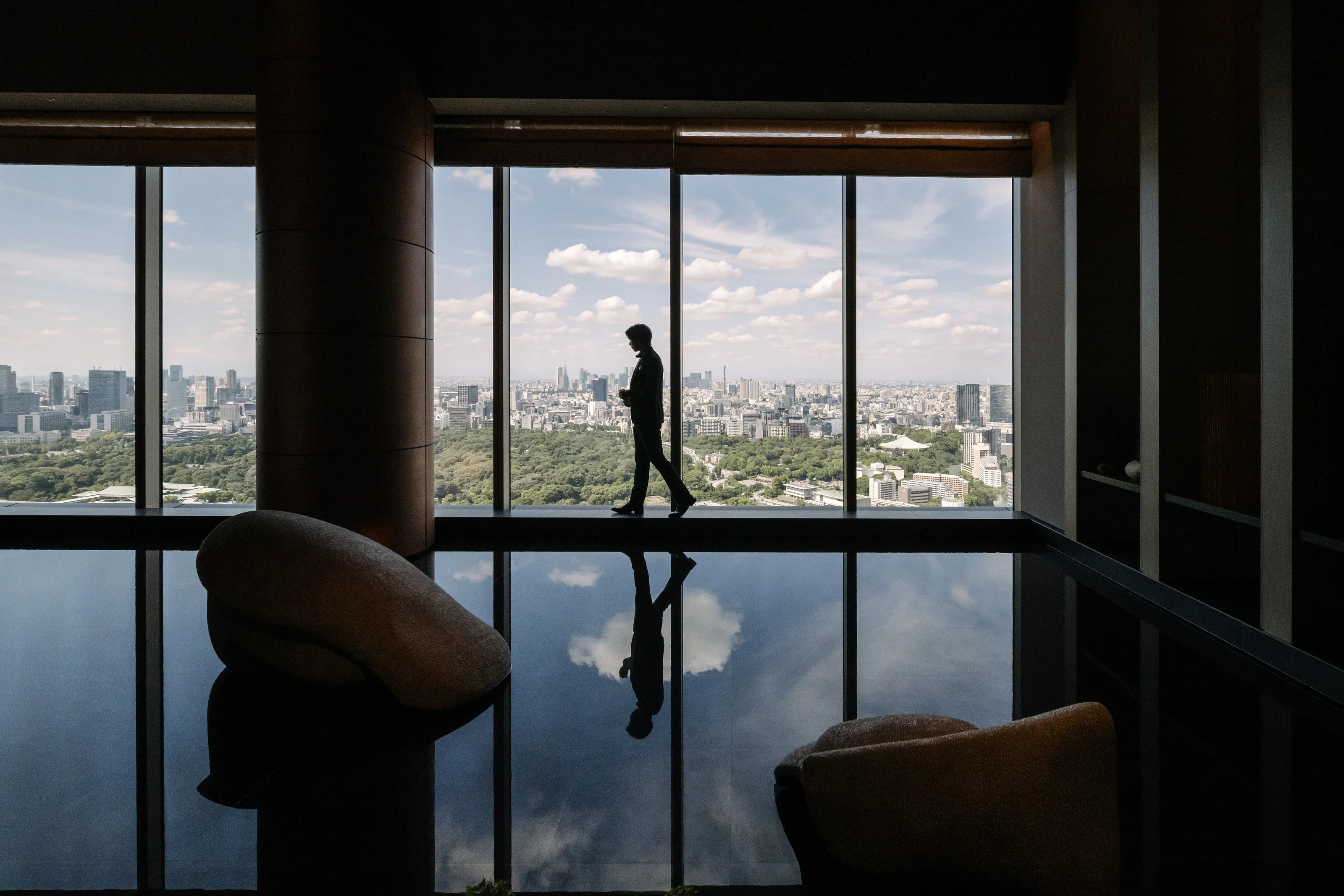 A figure walks past floor-to-ceiling windows high above Tokyo, the skyline and clouds reflected in the polished floor.