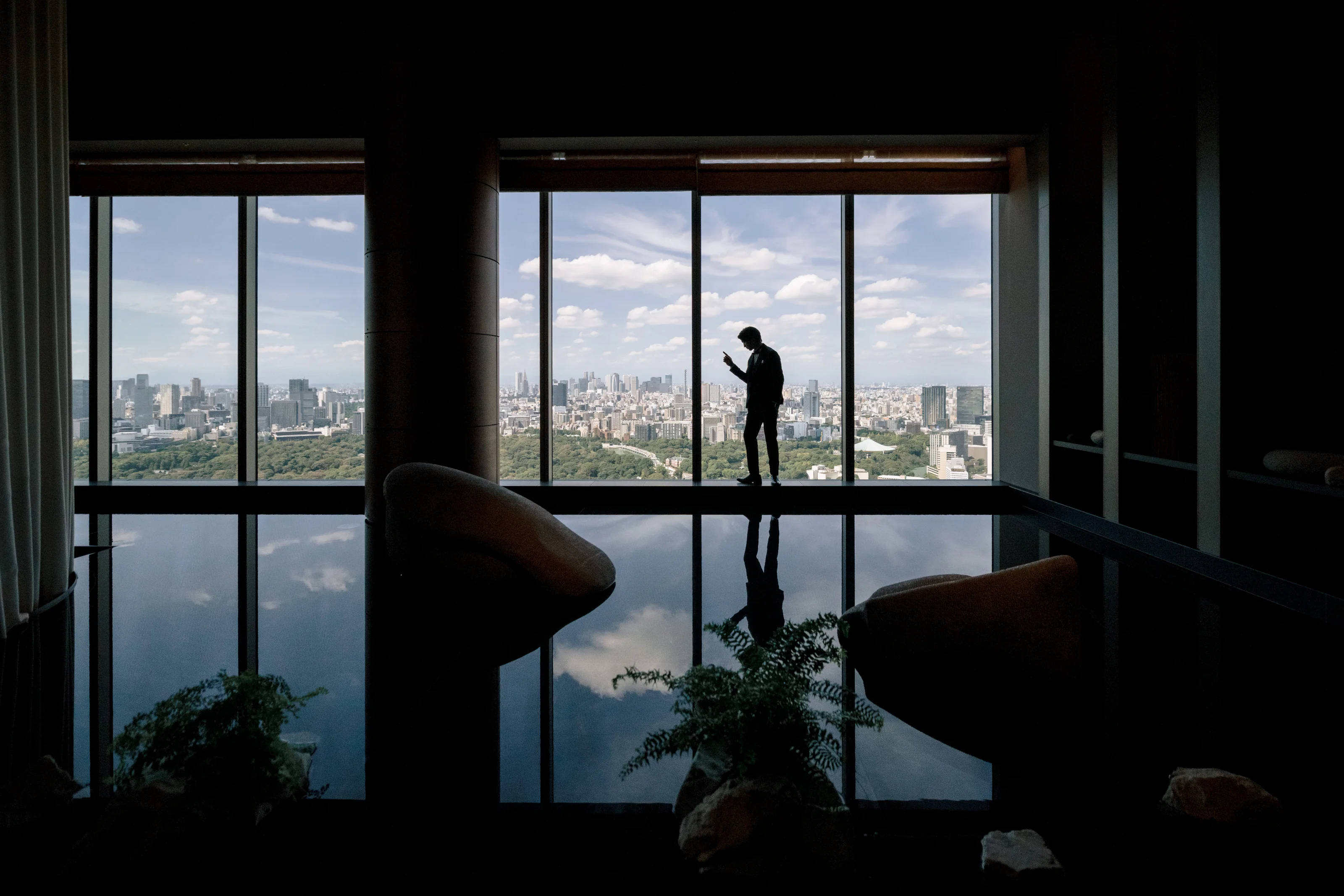 A lone figure in silhouette stands at a vast window high above Tokyo, the city and sky mirrored in the floor.