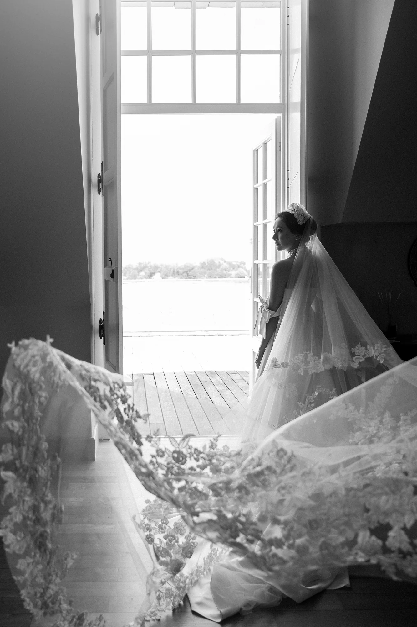 In black and white, the bride stands at a tall open door as her long lace veil spills across the floor, in France.