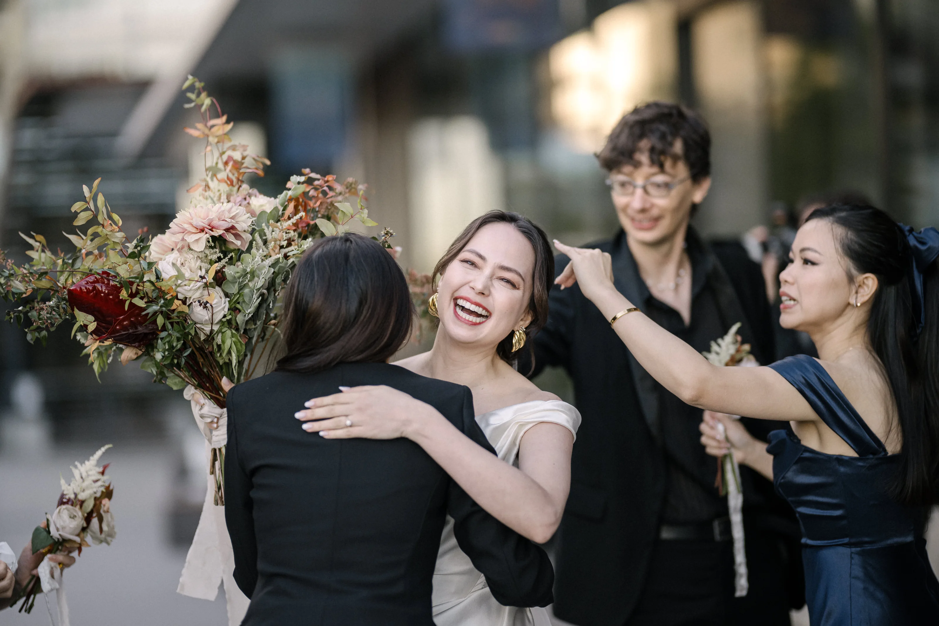Friends throw their arms around the bride, laughing, her bouquet of dried flowers in hand.