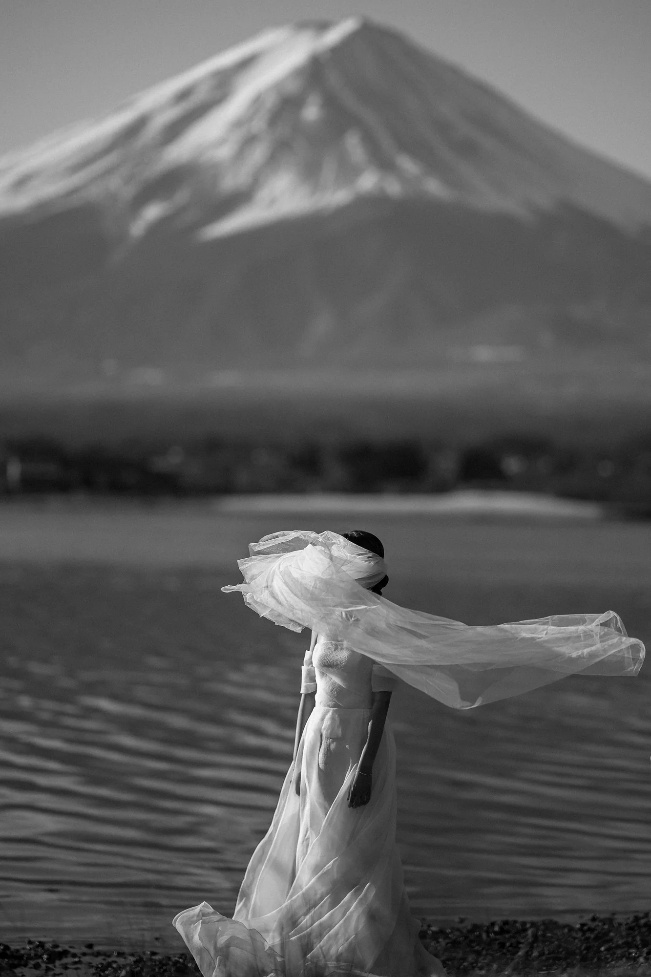 In black and white, a bride stands at the edge of Lake Kawaguchiko below Mt Fuji, her veil lifting in the wind.