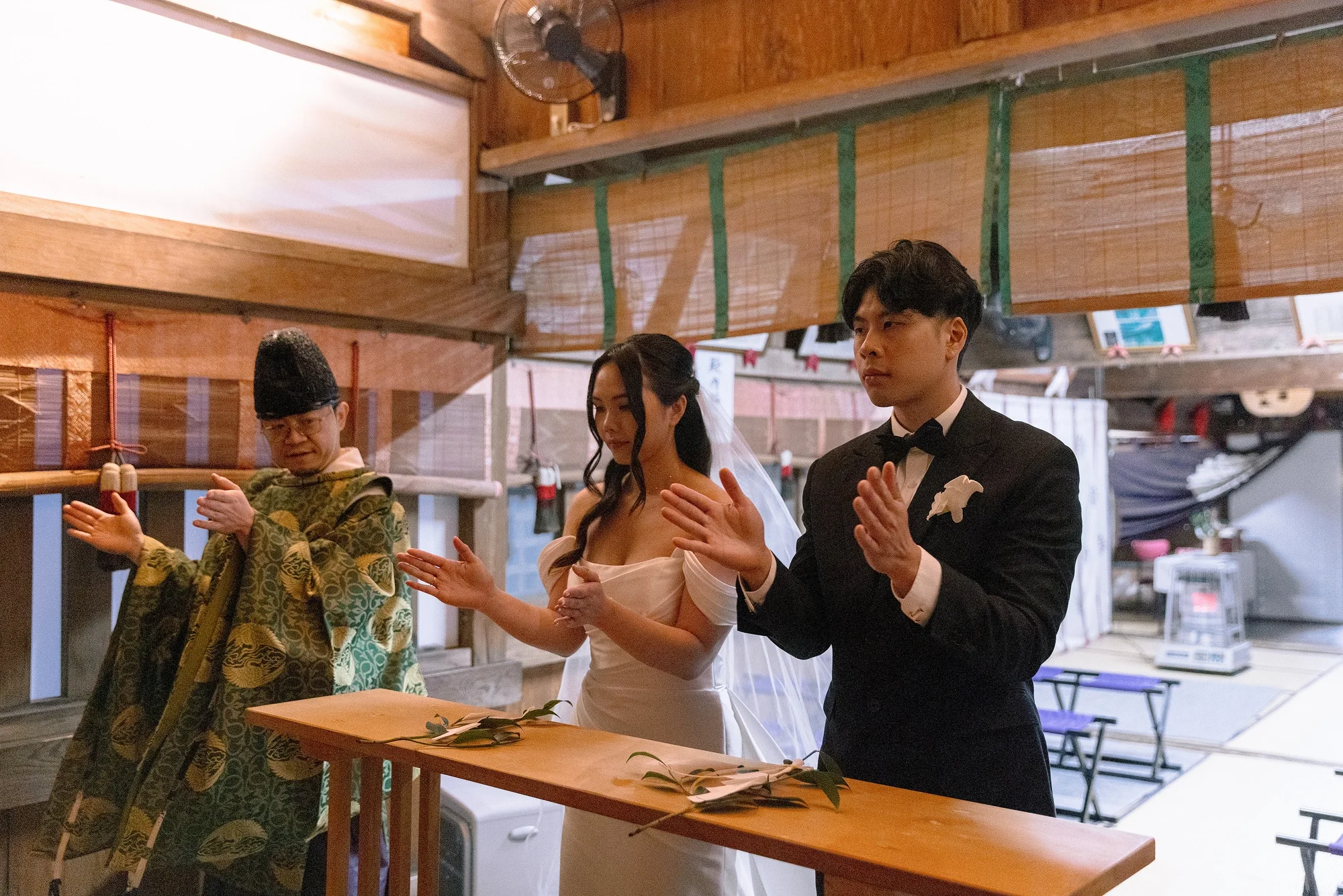 The bride in her gown and the groom clap with the priest at their Shinto blessing at Fuji Omuro Sengen Jinja.