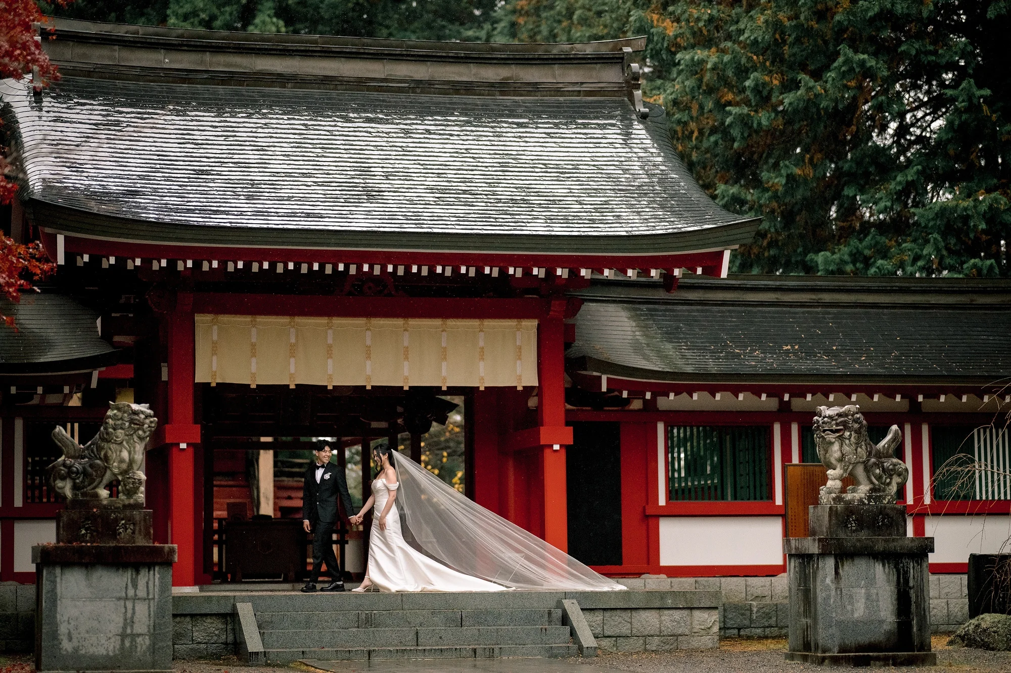 The couple walk out through the vermilion gate of Fuji Omuro Sengen Jinja, her long veil trailing on the wet stone.