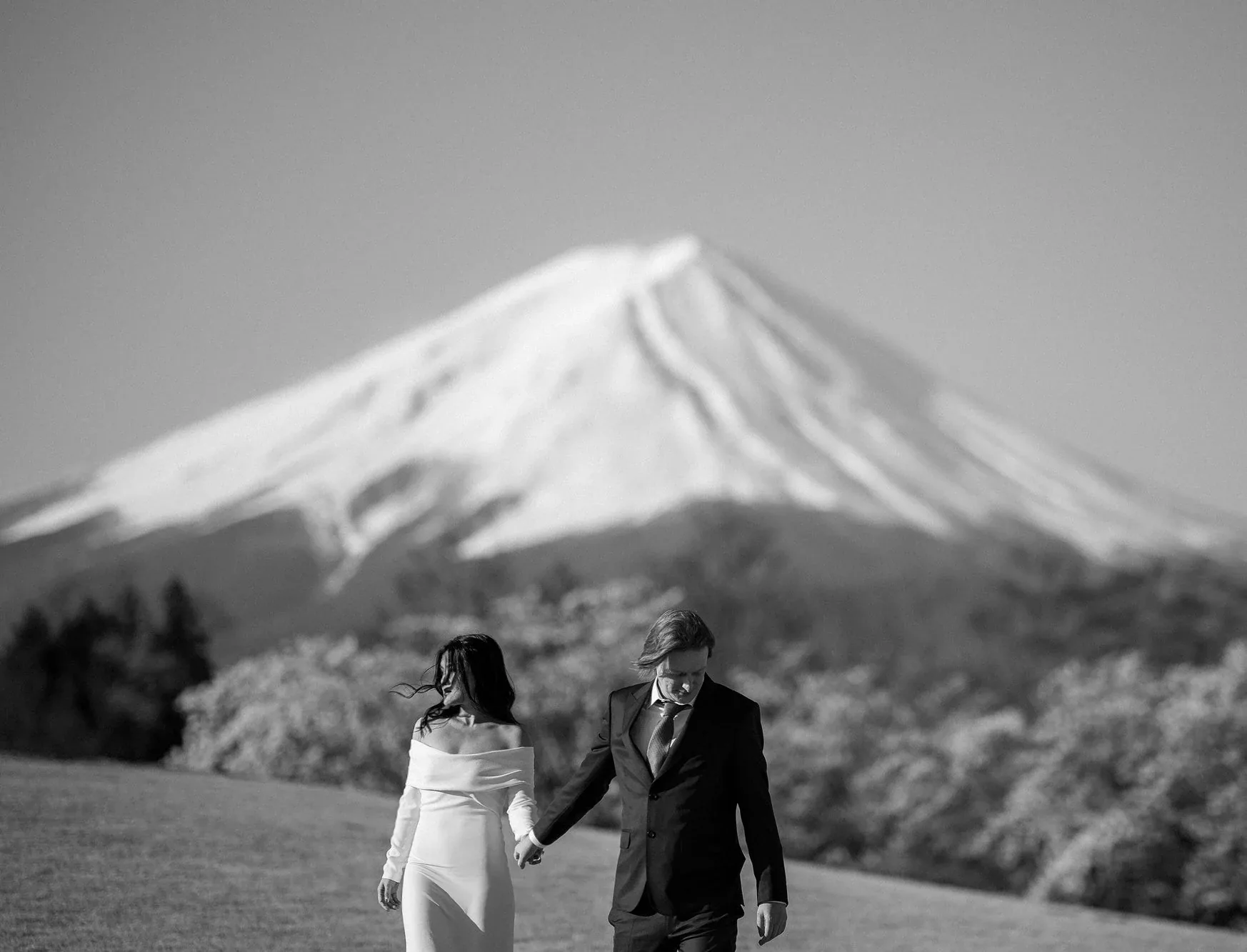 In black and white, a couple walk hand in hand beneath Mt Fuji, wind in the bride's hair.