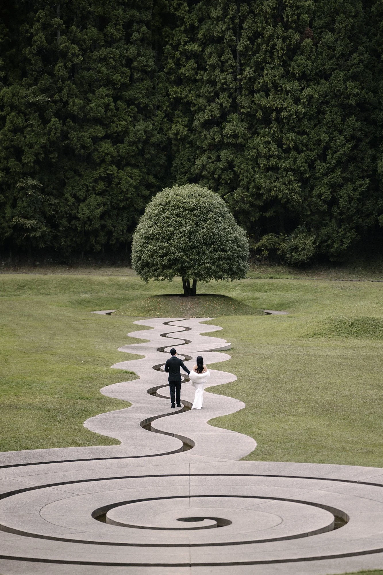 A couple walk along a sculptural path toward a tree, with forest beyond.