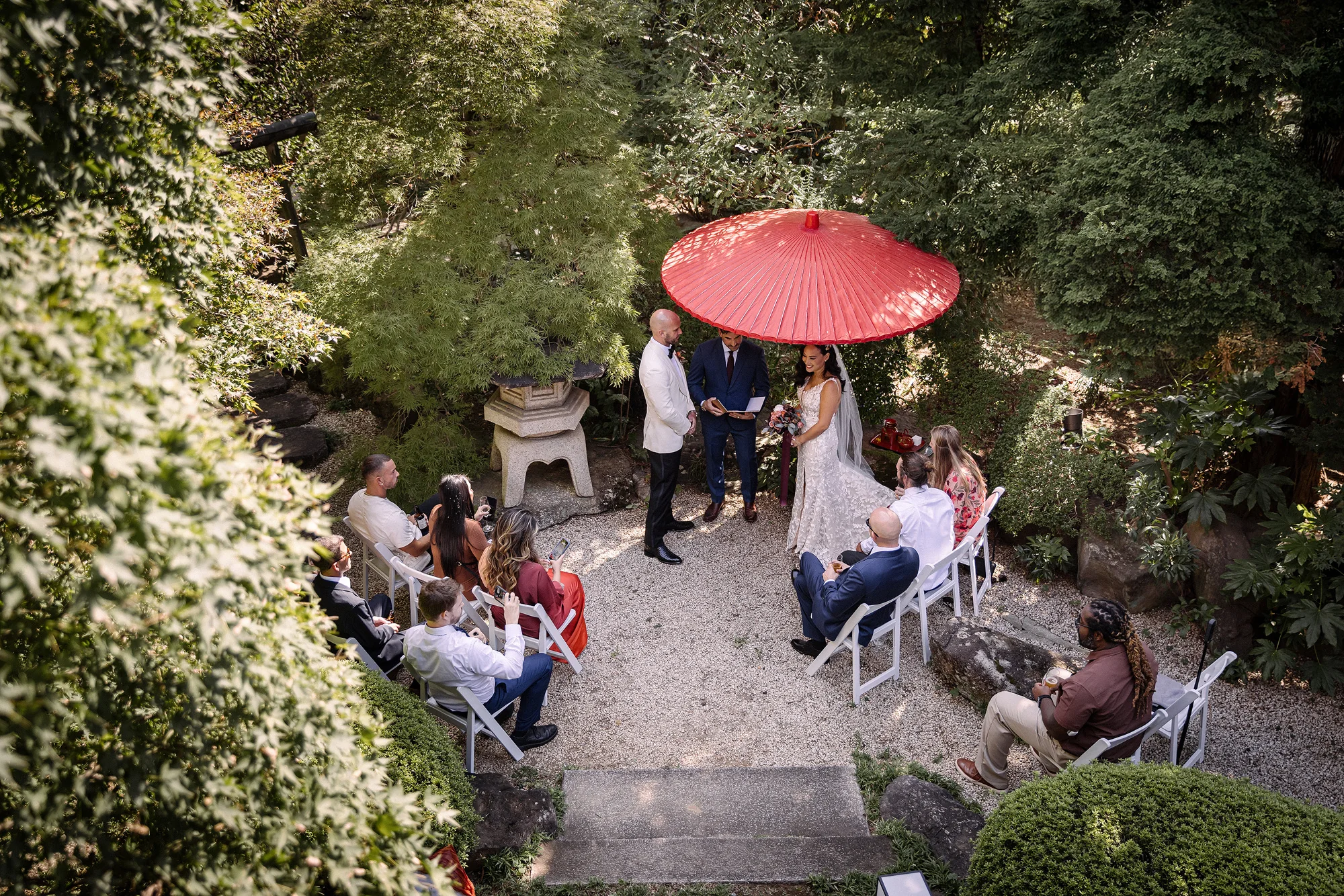 Guests gather in a gravel garden for an intimate ceremony under a red parasol.