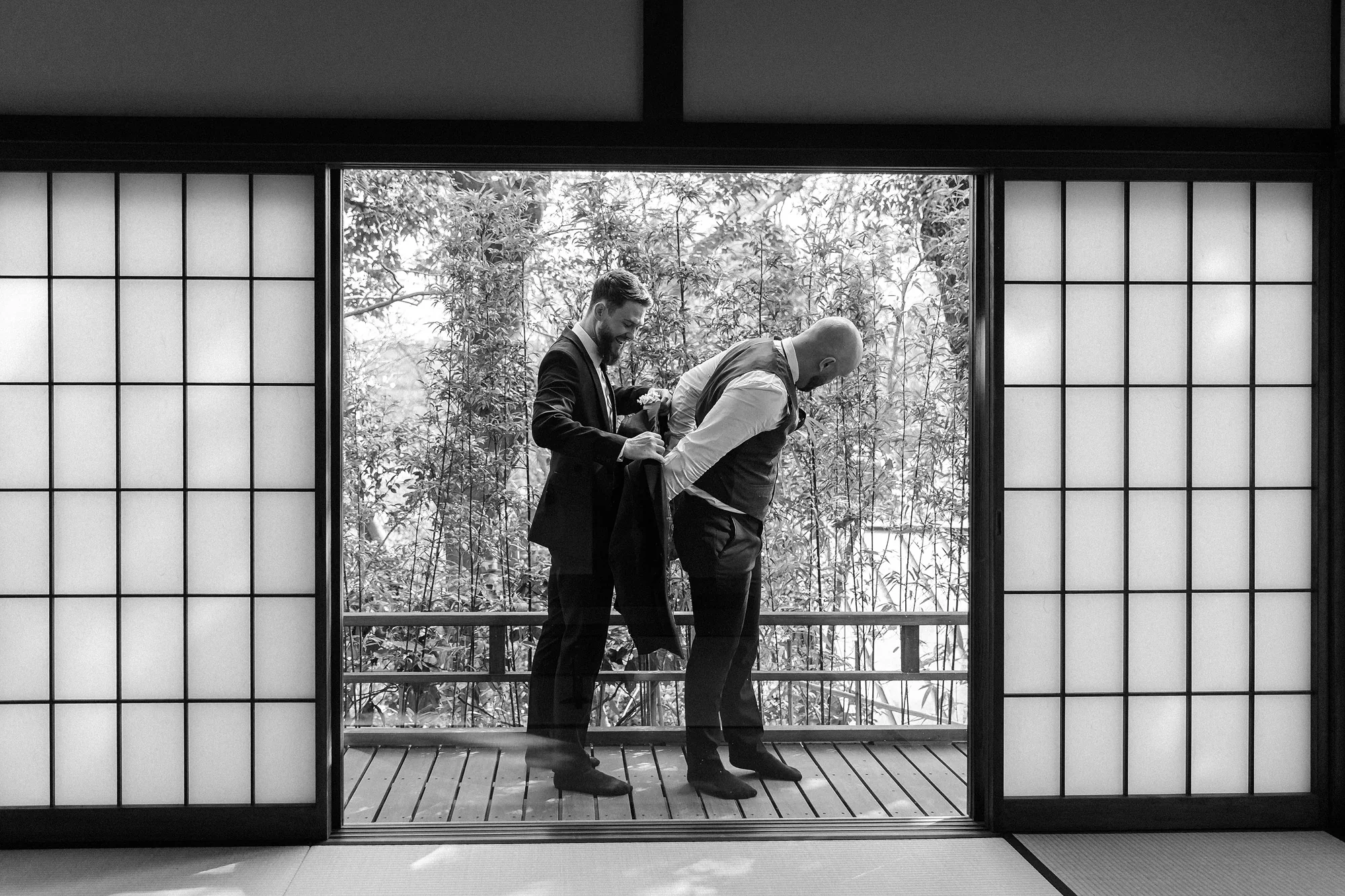 In black and white, the best man helps the groom into his jacket between open shoji doors in Hakone, bamboo beyond.