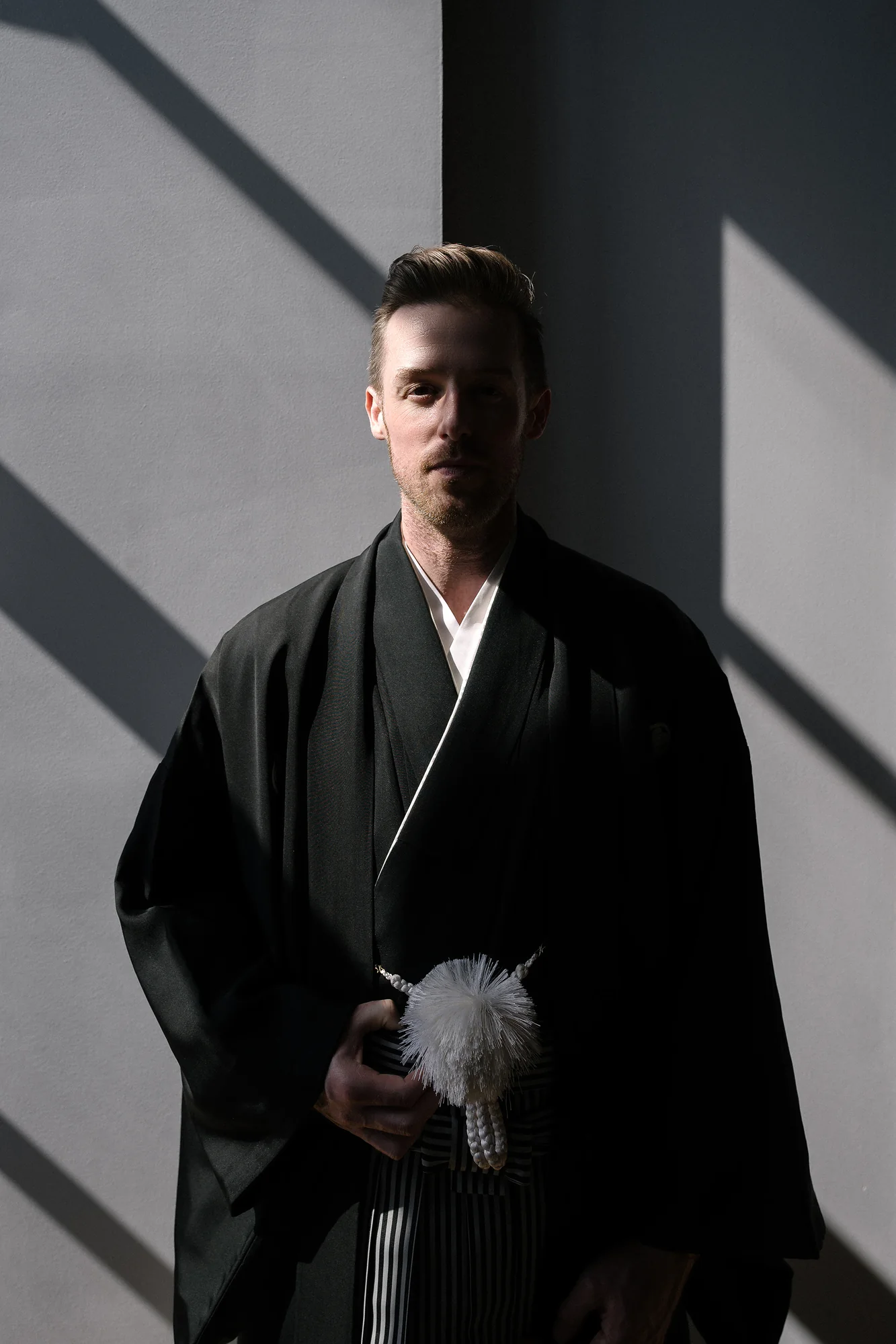 The groom in a black montsuki and striped hakama stands half in shadow against a pale wall crossed by bars of light.