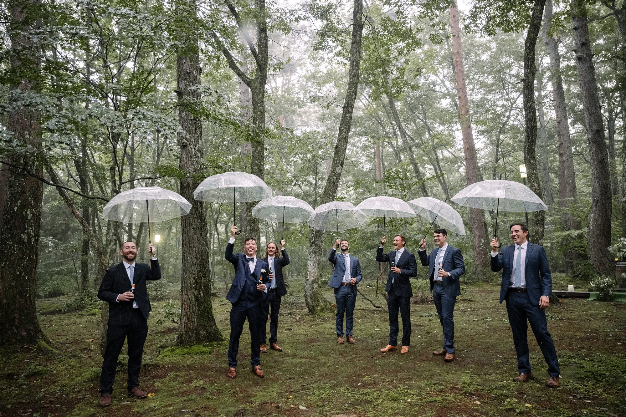 Groomsmen under clear umbrellas laugh and dance in a rainy forest.