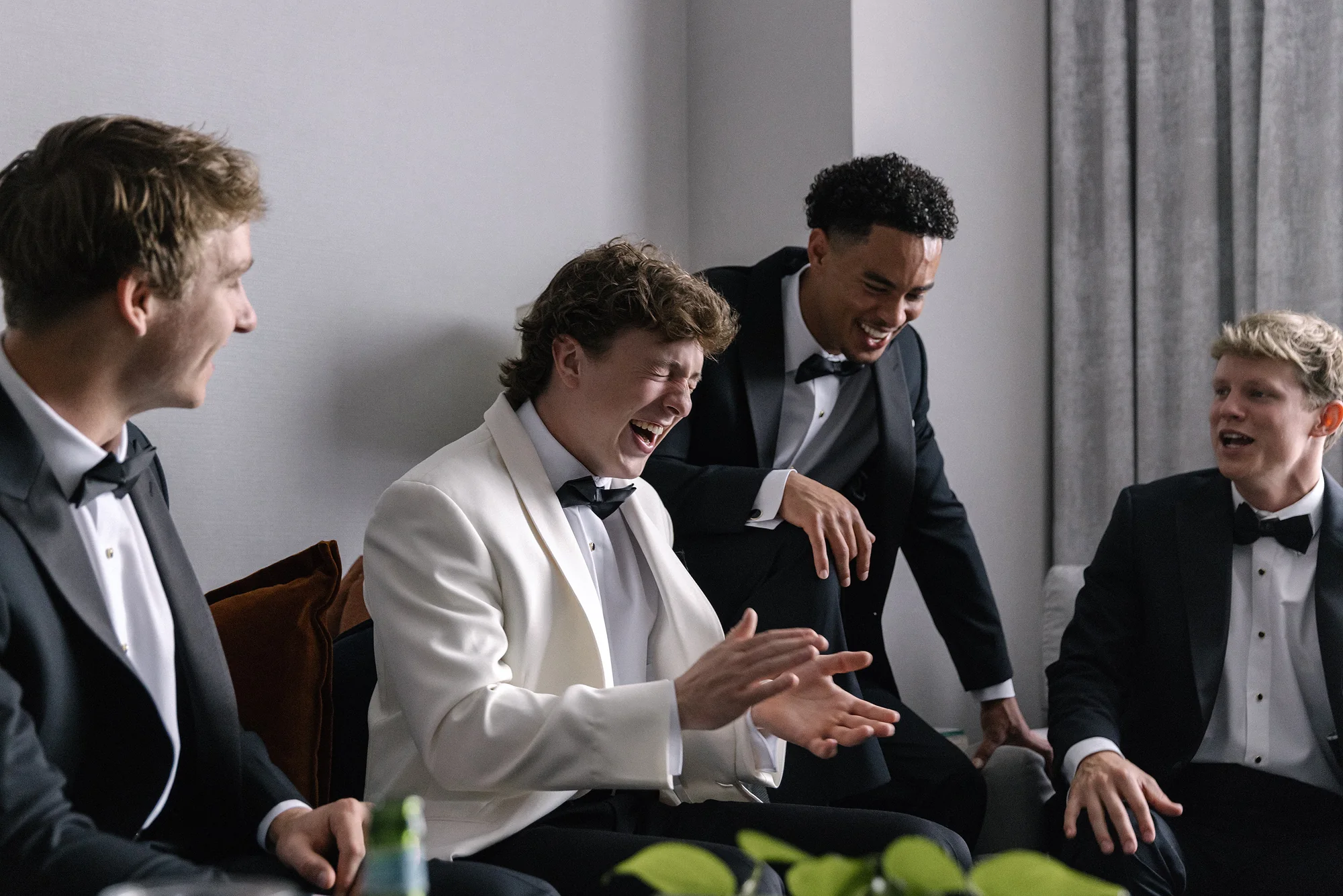 The groom in a white dinner jacket laughs with his groomsmen on a sofa as they get ready.