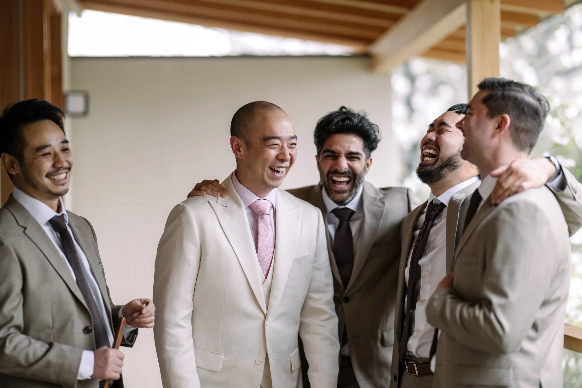 The groom and his groomsmen in pale suits laugh together on a wooden veranda.