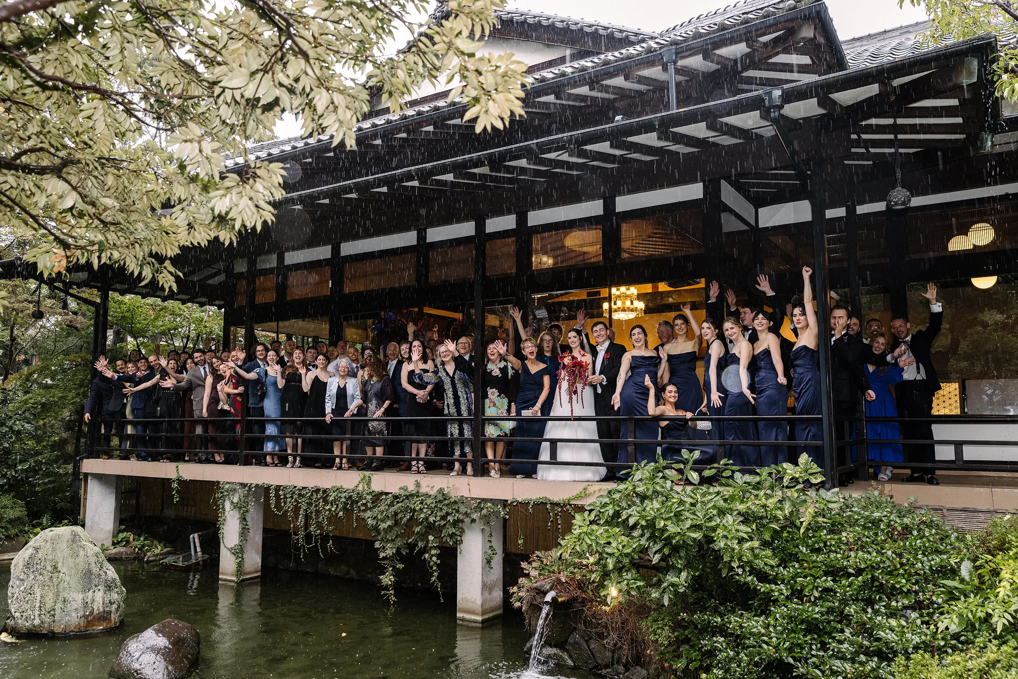 Wedding guests crowd the veranda of a timber hall over a garden pond, waving and cheering.