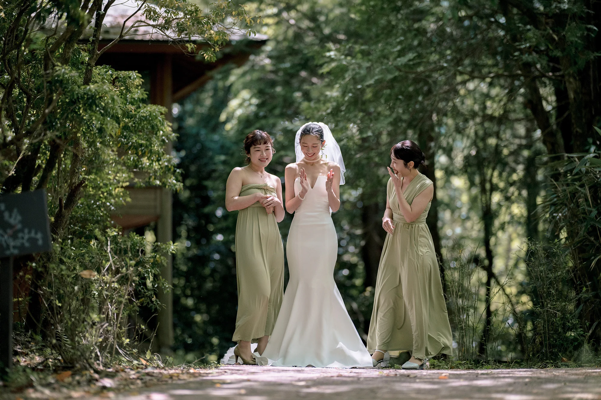 The bride walks between two bridesmaids in sage green along a forest path in Hakone.