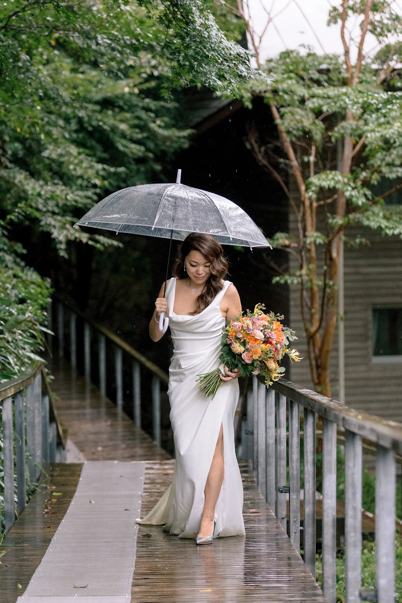 The bride crosses a wet wooden bridge under a clear umbrella, bouquet in hand, on her way to the ceremony in Hakone.