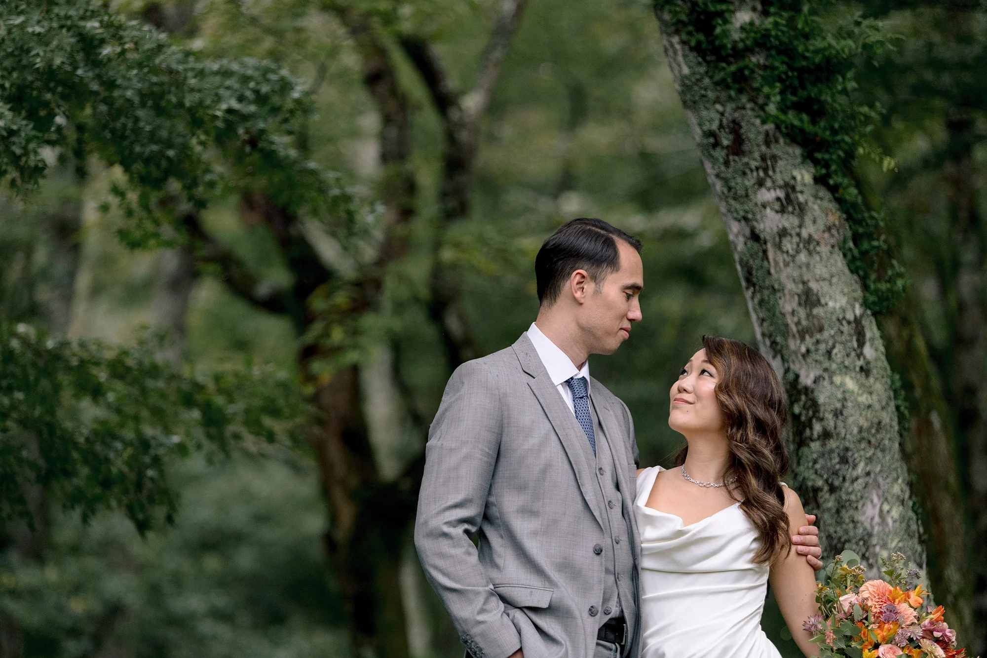 The groom in a gray suit and the bride in an off-the-shoulder gown look at each other beneath tall trees in Hakone.