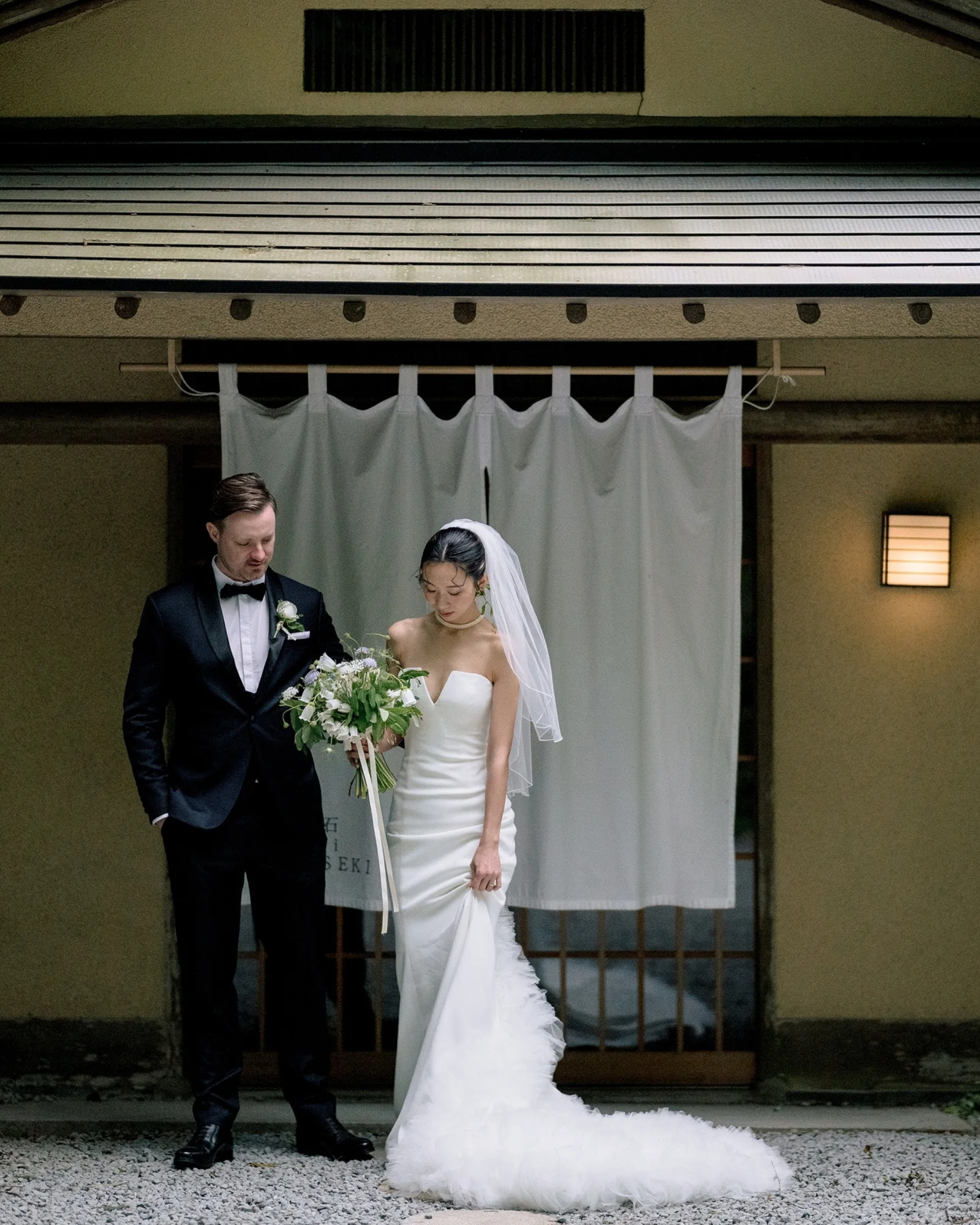 The bride and groom stand together before a white noren curtain at a doorway in Hakone.