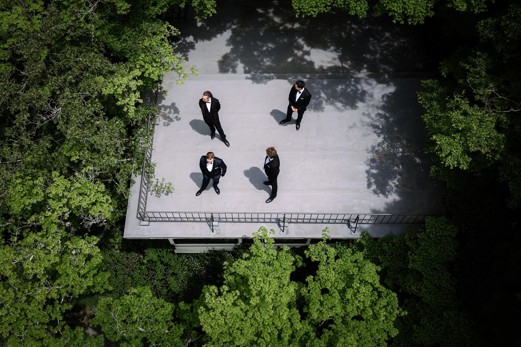 Seen from above, four groomsmen in black tie stand on a white deck in the forest canopy in Hakone.