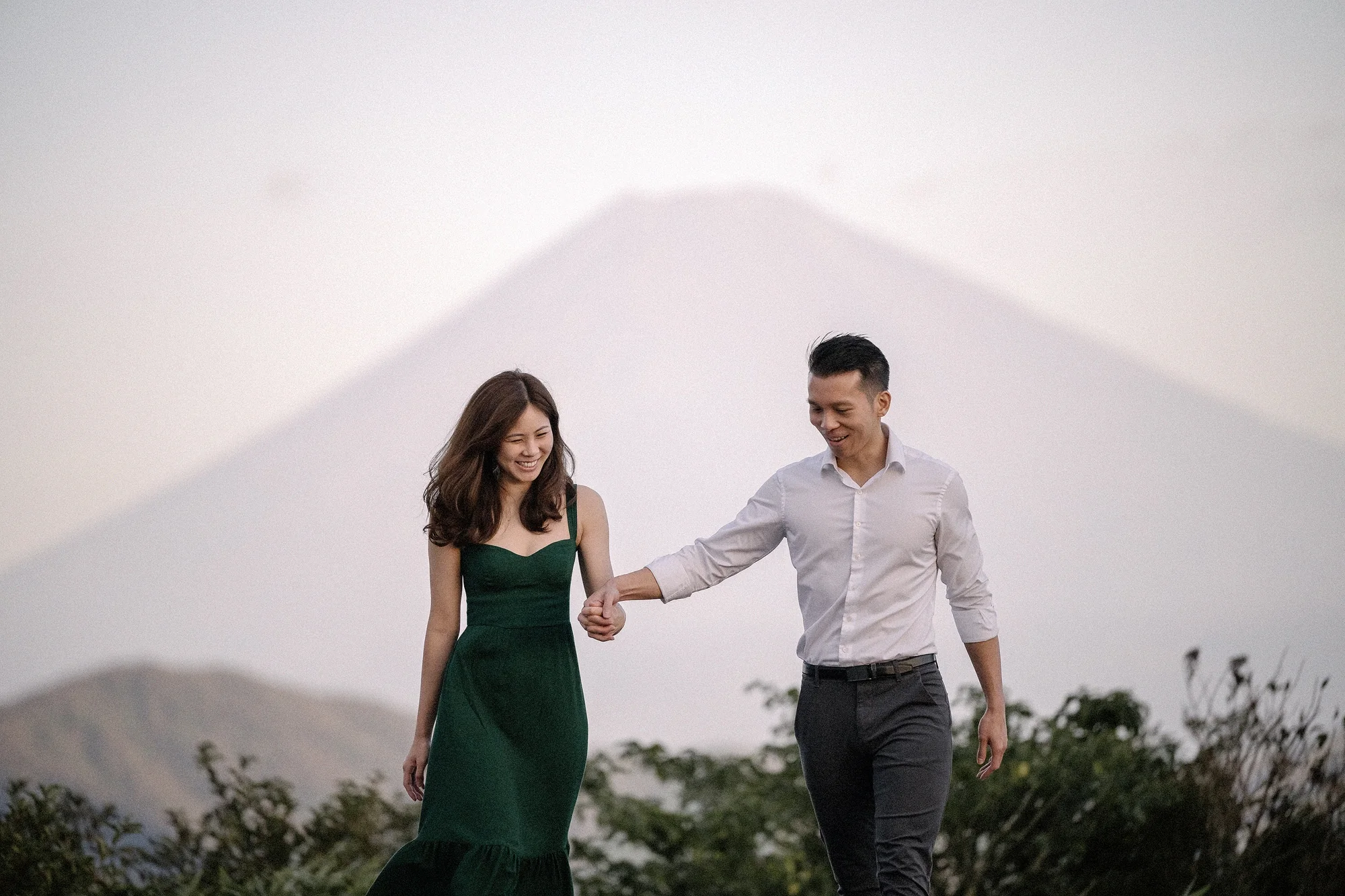 A couple walk hand in hand on a Hakone hilltop, Mt Fuji pale in the morning sky behind them.